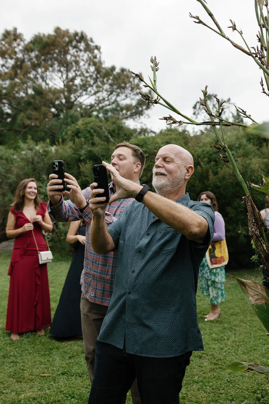 Older man and young man taking photos with smartphones at a garden party, with two women smiling in the background. ocracoke outerbanks wedding film photographer