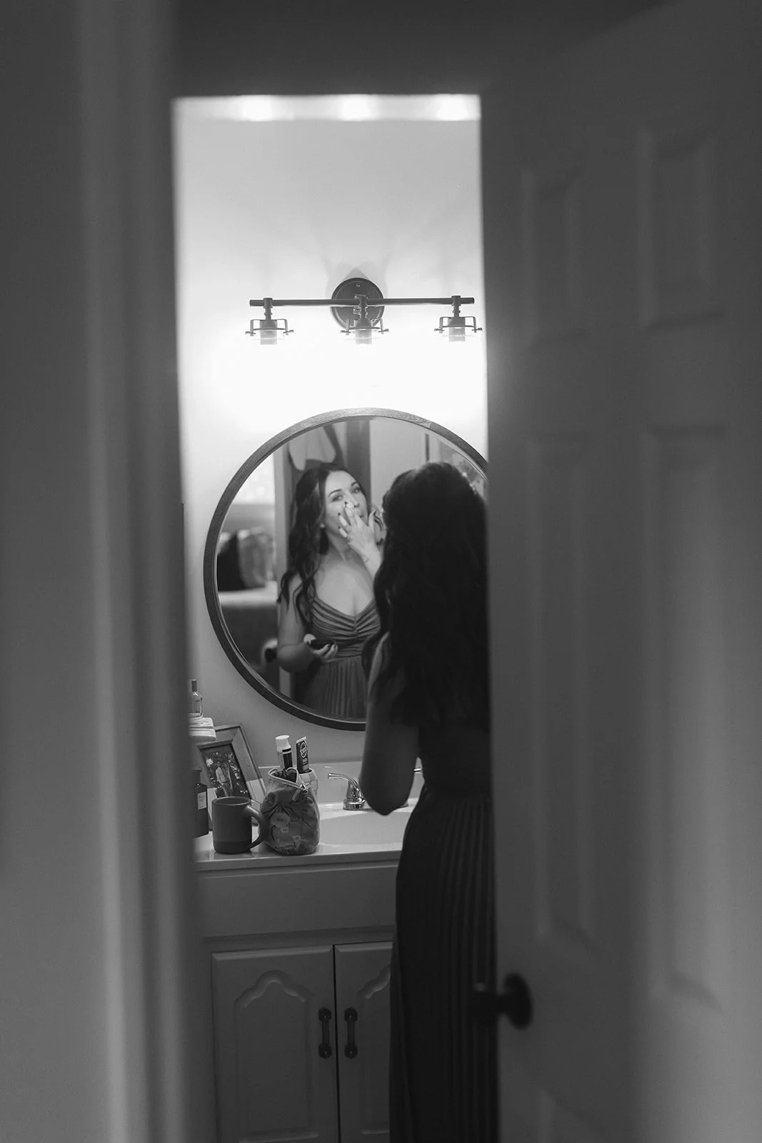 A woman looking in a bathroom mirror, touching her face, while applying makeup, with her reflection visible, and personal items on the bathroom counter. Roanoke Virginia Wedding Photographer.