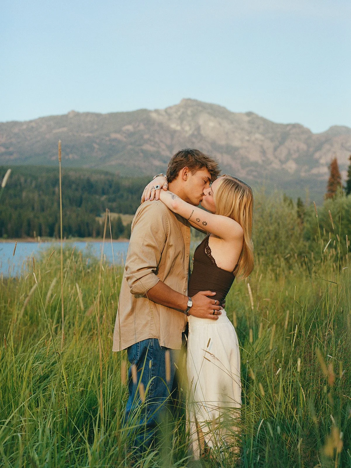A young couple sharing a kiss in a grassy field with mountains and a lake in the background. bozeman wedding photographer