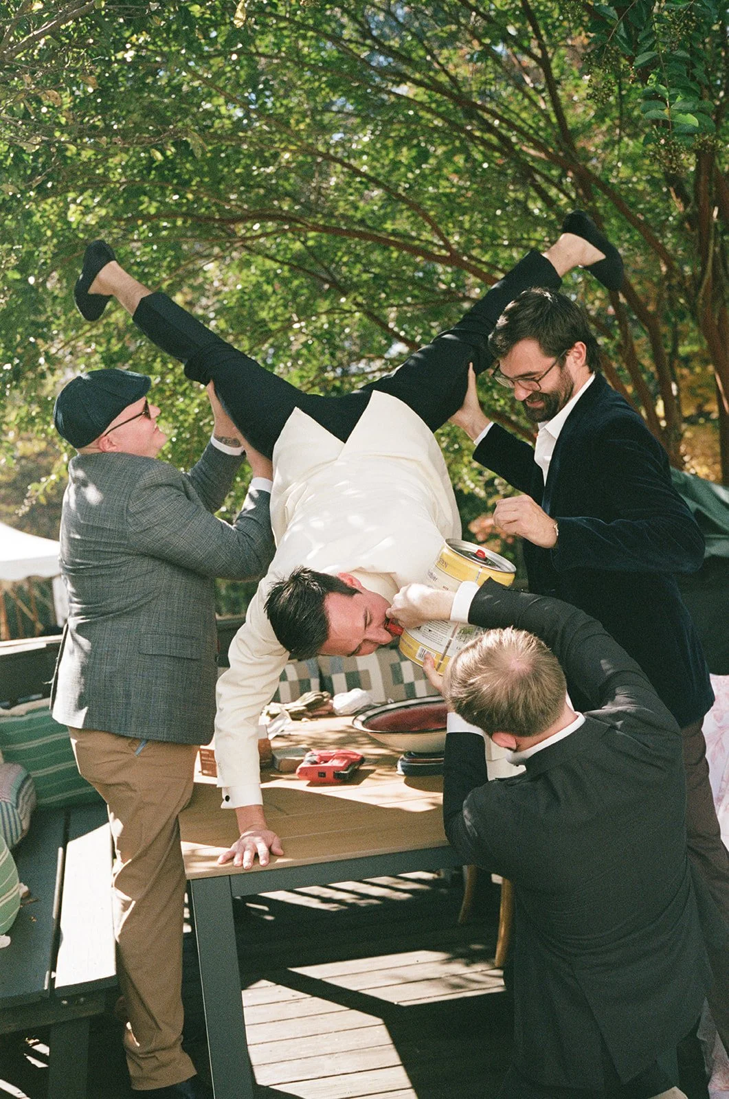 A group of five men are playfully lifting a man in white shirt over a table outdoors under trees during daytime. Charlottesville Virginia Wedding Film Photographer