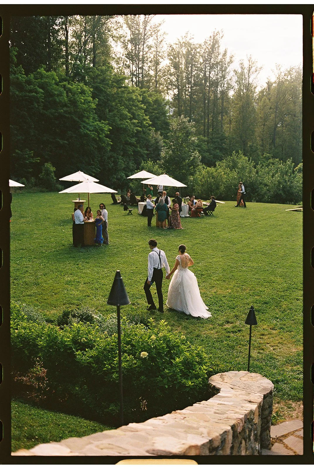 A bride and groom walk hand in hand on a grassy lawn during a wedding reception in the late afternoon or early evening. Guests are seated under white umbrellas and standing around tables, enjoying the outdoor celebration surrounded by lush greenery and trees.