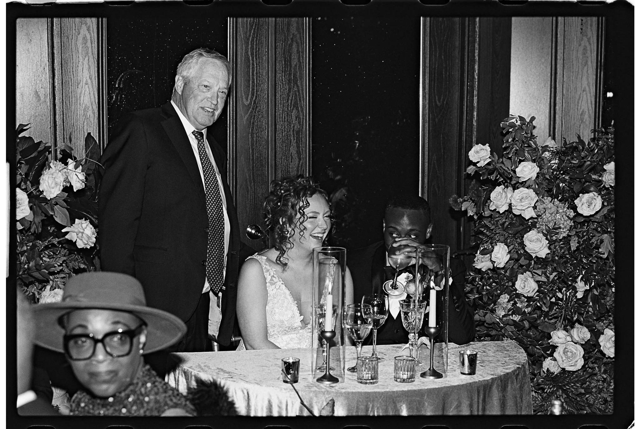 Black and white photo of a wedding reception with three people gathered around a table. A man in a suit stands beside a sitting bride and groom, who are sharing a moment of laughter. The table is set with glasses and candles, adorned with floral arra
