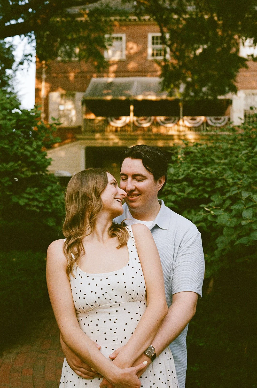 A happy couple standing close together outdoors, smiling and looking into each other's eyes with a house and greenery in the background. Old City Philadelphia engagement shoot