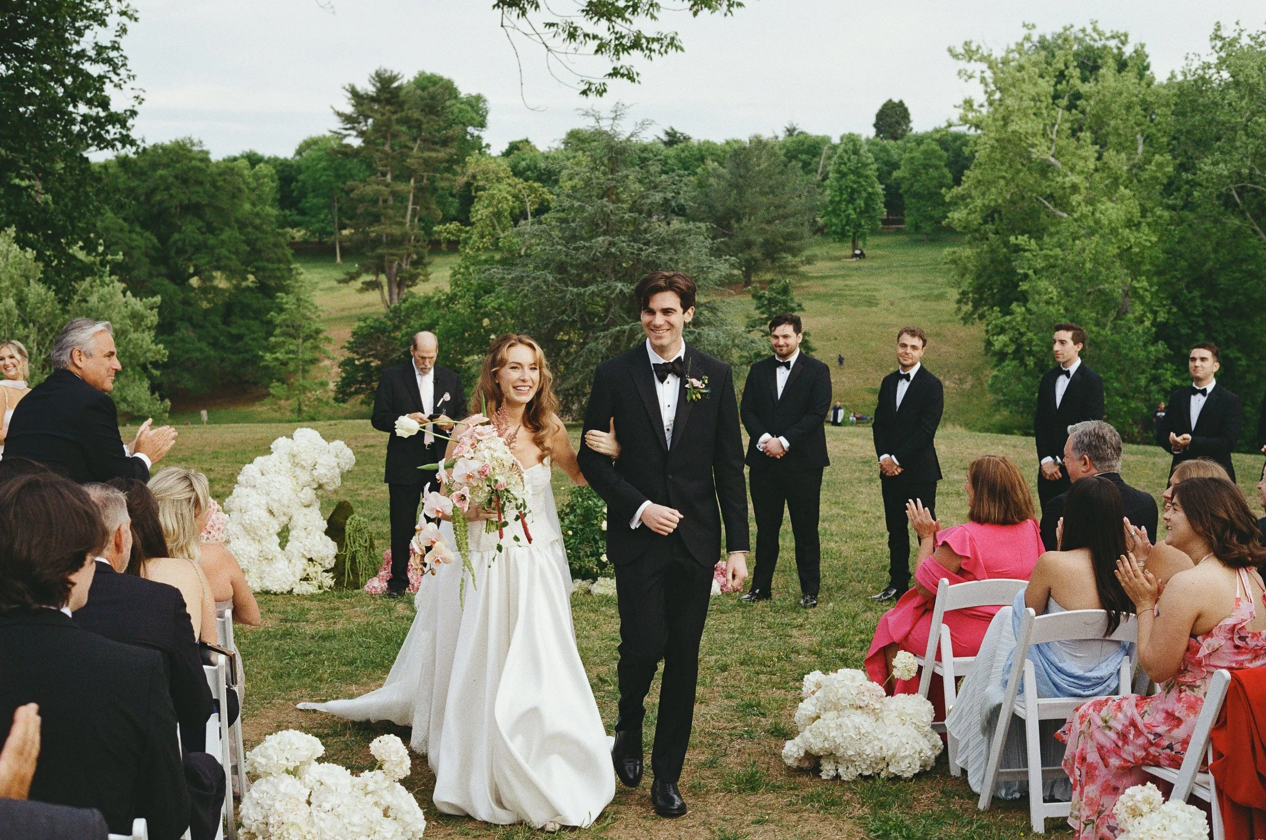 A wedding ceremony outdoors with a bride and groom walking down the aisle, surrounded by seated guests and groomsmen in black tuxedos, set in a lush green park. richmond virginia wedding photographer