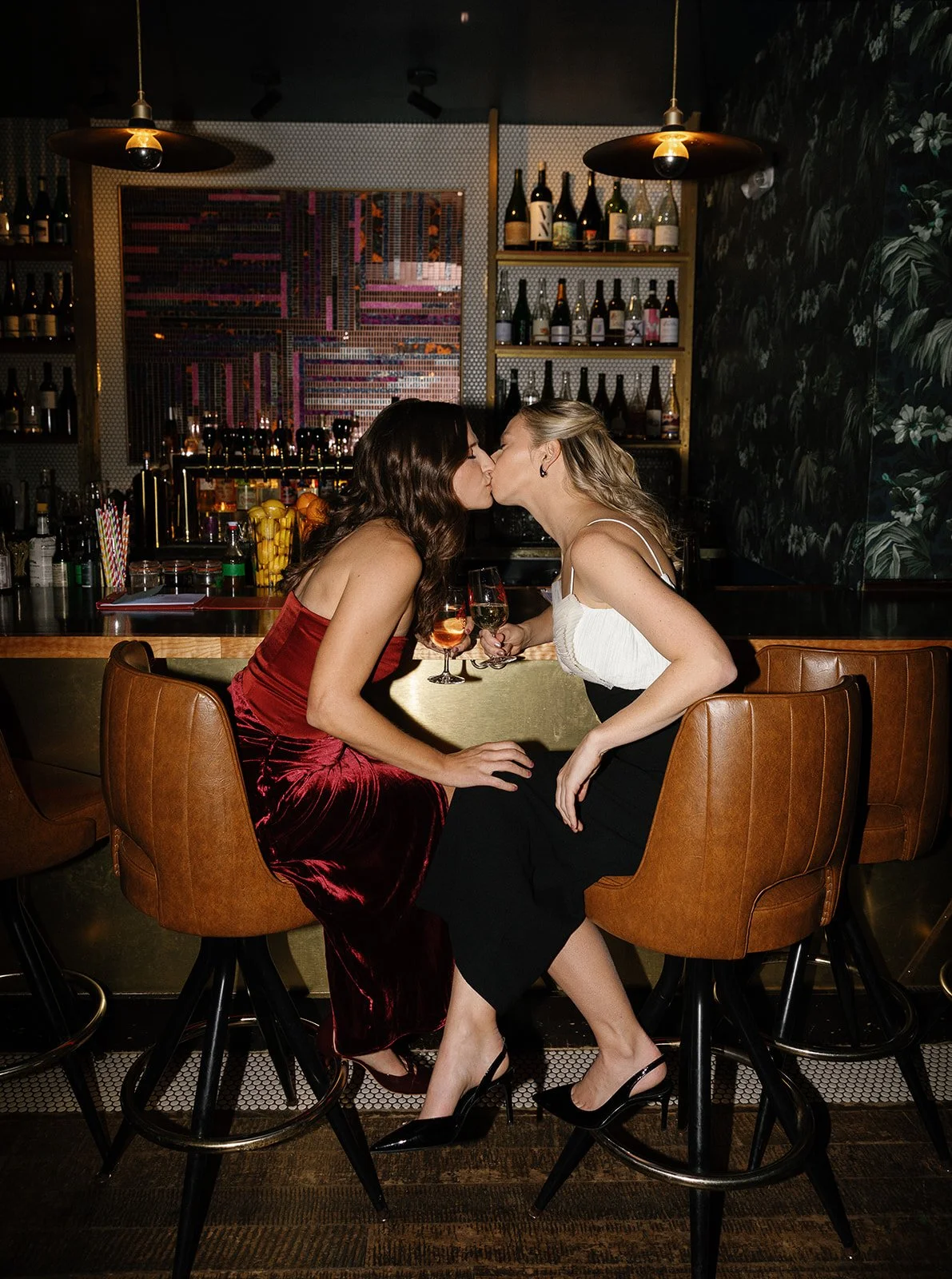 Two women are sitting in a bar, sharing a kiss while holding glasses of wine, with a bar counter and bottles in the background. DC Wedding film photographer no kisses bar