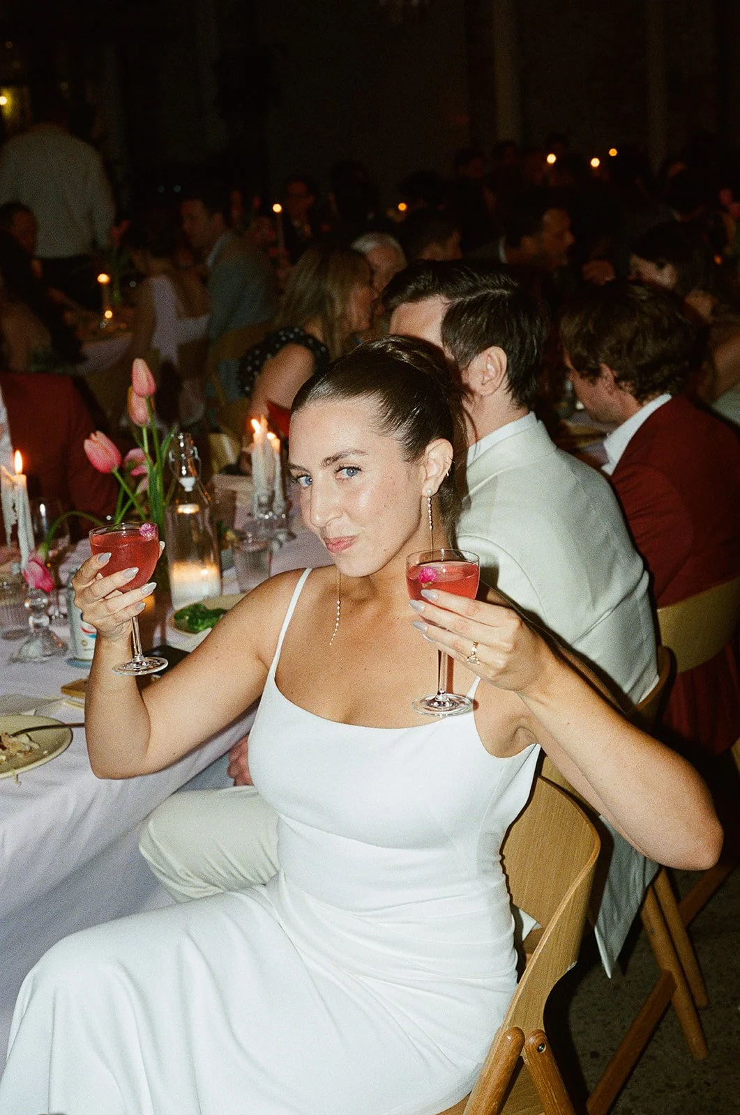 A woman in a white dress holding a pink cocktail at a formal dinner event with many guests and candlelit table. brooklyn new york wedding film photographer
