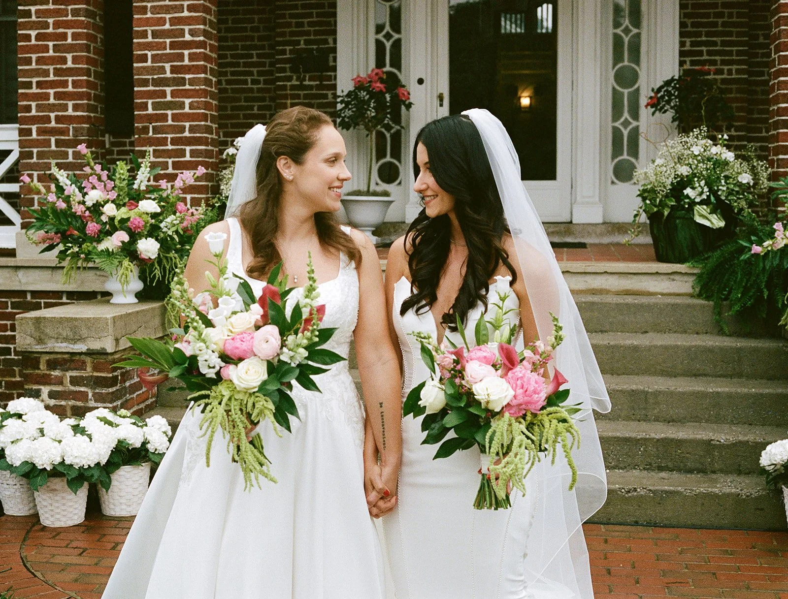 Two women in wedding dresses holding hands and bouquets, standing on steps outside a brick building decorated with flowers. roanoke wedding film photographer virginia