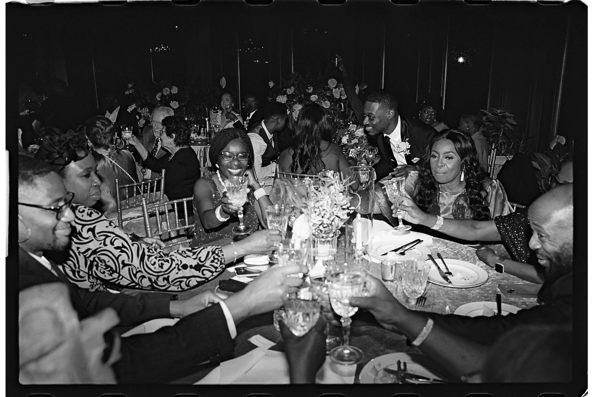 A black-and-white photo of a group of people sitting around a table, raising glasses in a toast at a formal event. They are dressed in formal attire, and there are plates and glasses on the table, along with floral centerpieces in the background.