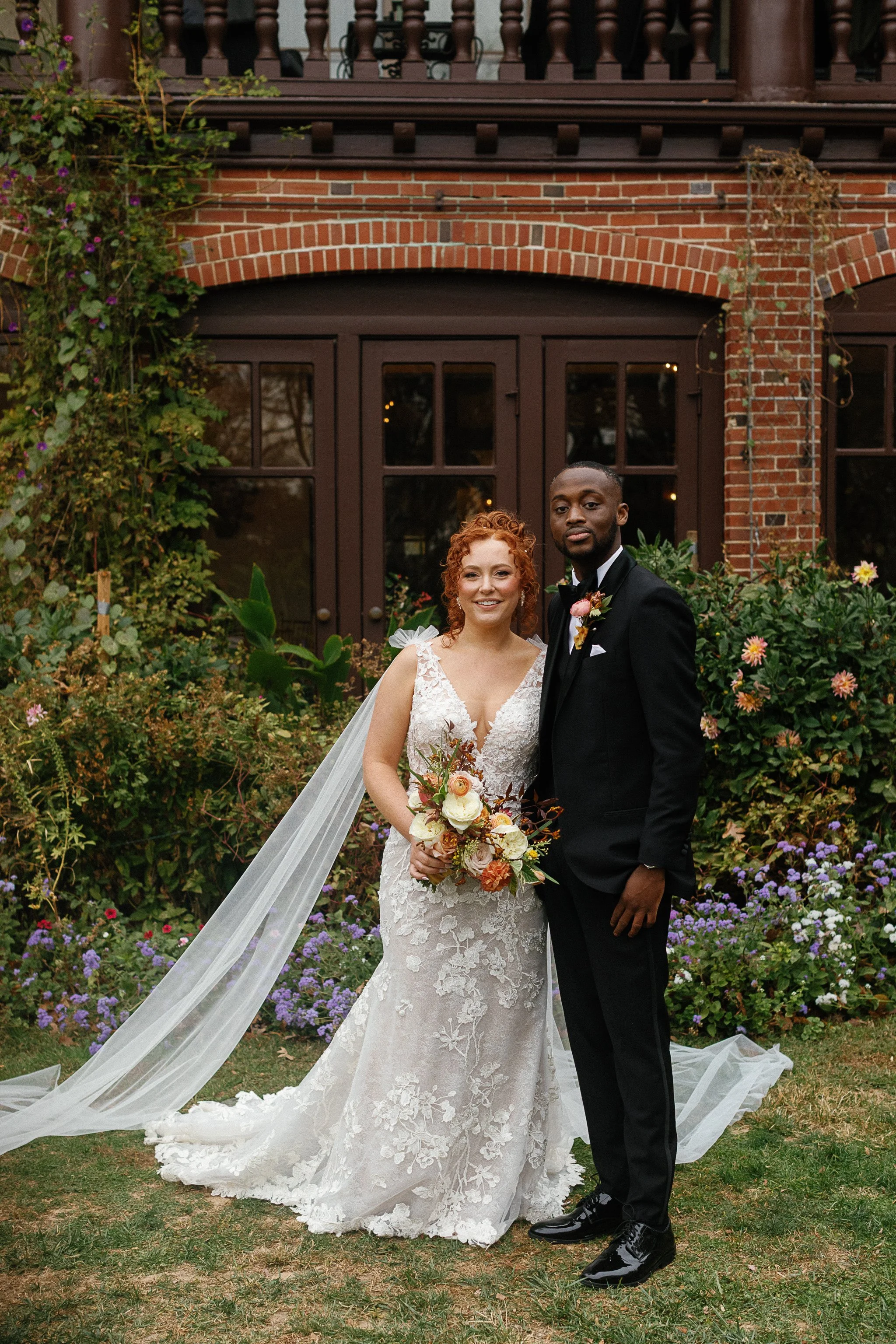 Bride and groom posing in front of a brick building, bride in a lace wedding dress holding a bouquet, groom in a black tuxedo, surrounded by greenery and flowers. gramercy mansion maryland wedding