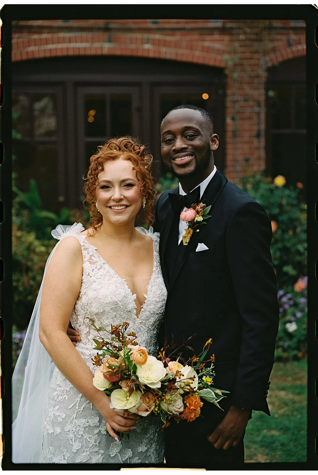 A newlywed couple stands together outdoors in front of a brick building. The bride has curly red hair and is wearing a white lace wedding dress, holding a bouquet of flowers. The groom has short black hair and a beard, wearing a black tuxedo with a b