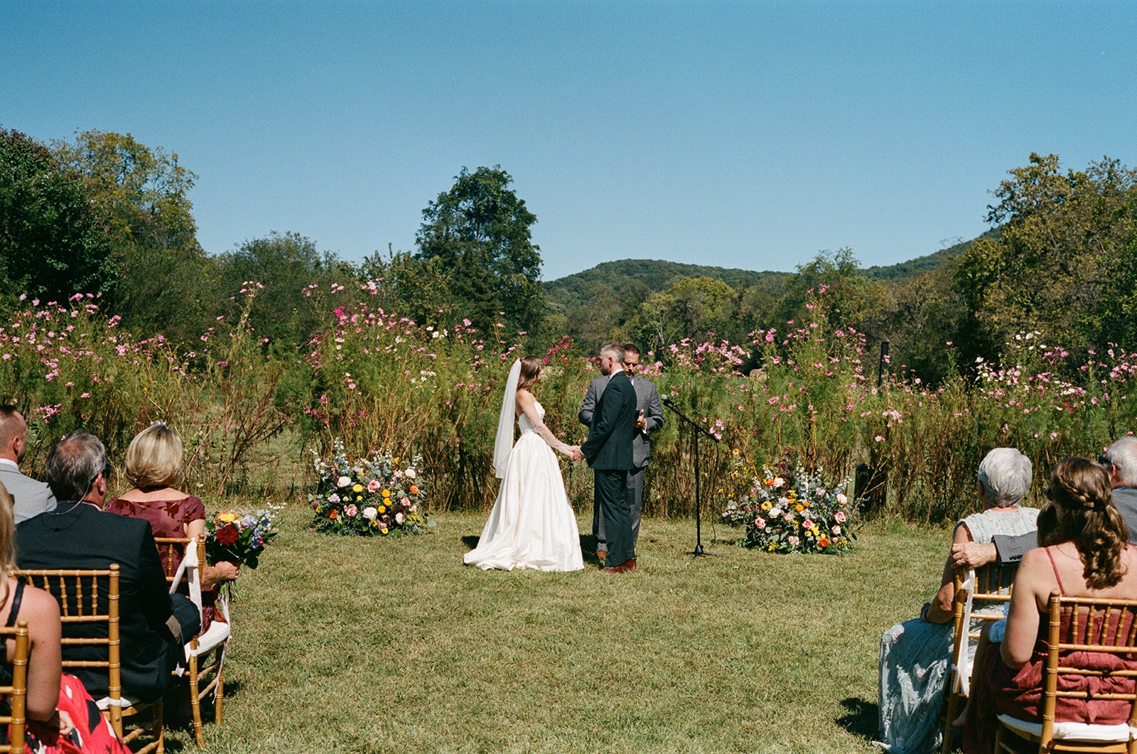 A wedding ceremony outdoors in a natural setting with mountains and trees in the background. The bride and groom are standing hand in hand, exchanging vows, with the officiant behind them. Guests are seated on either side, watching the ceremony, with some holding bouquets of flowers. The area is decorated with colorful floral arrangements.