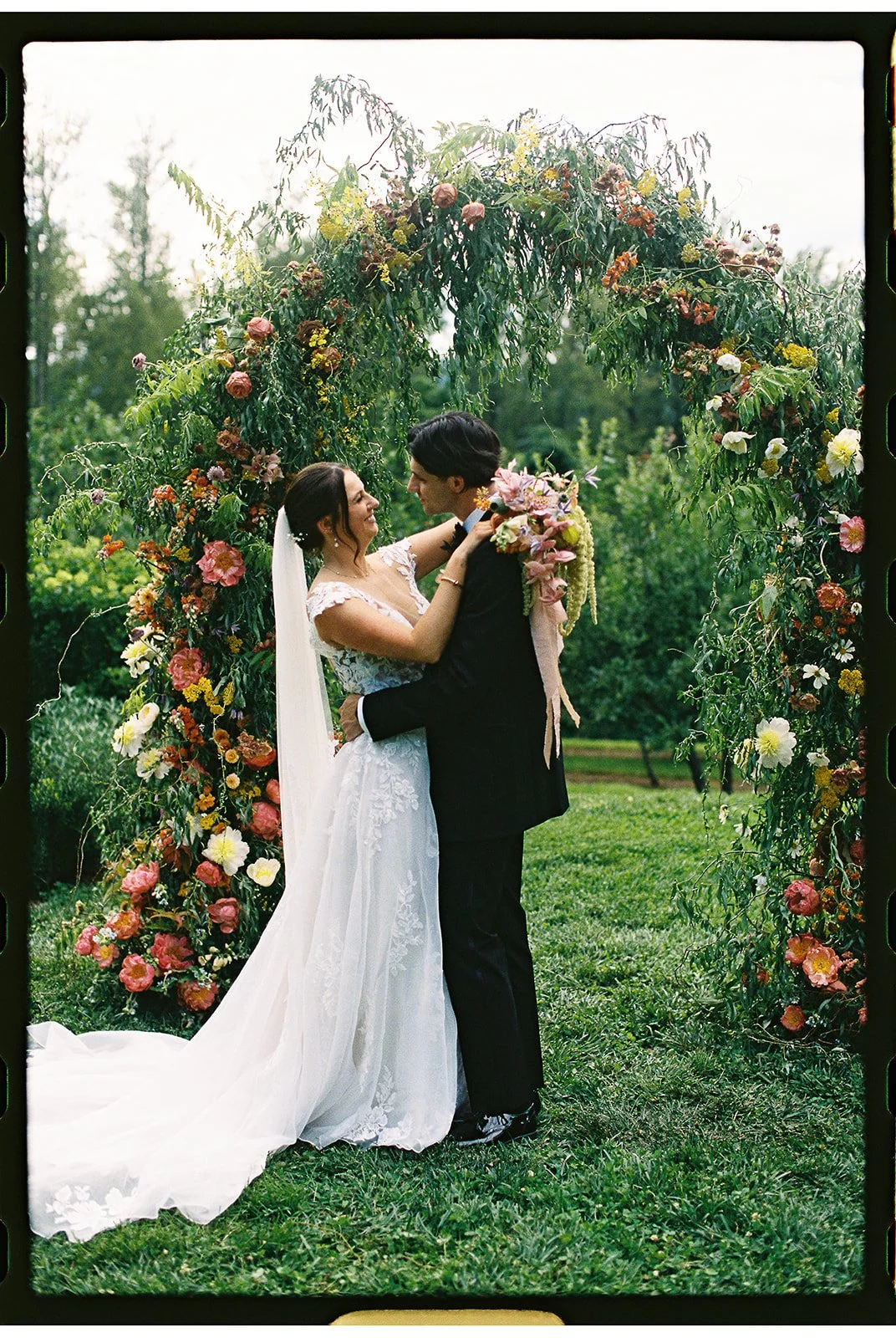 A bride and groom stand closely together under a floral arch on their wedding day outdoors, looking into each other's eyes. The bride is in a white lace wedding gown and veil, and the groom is in a black tuxedo with a large bouquet. The scene is set 