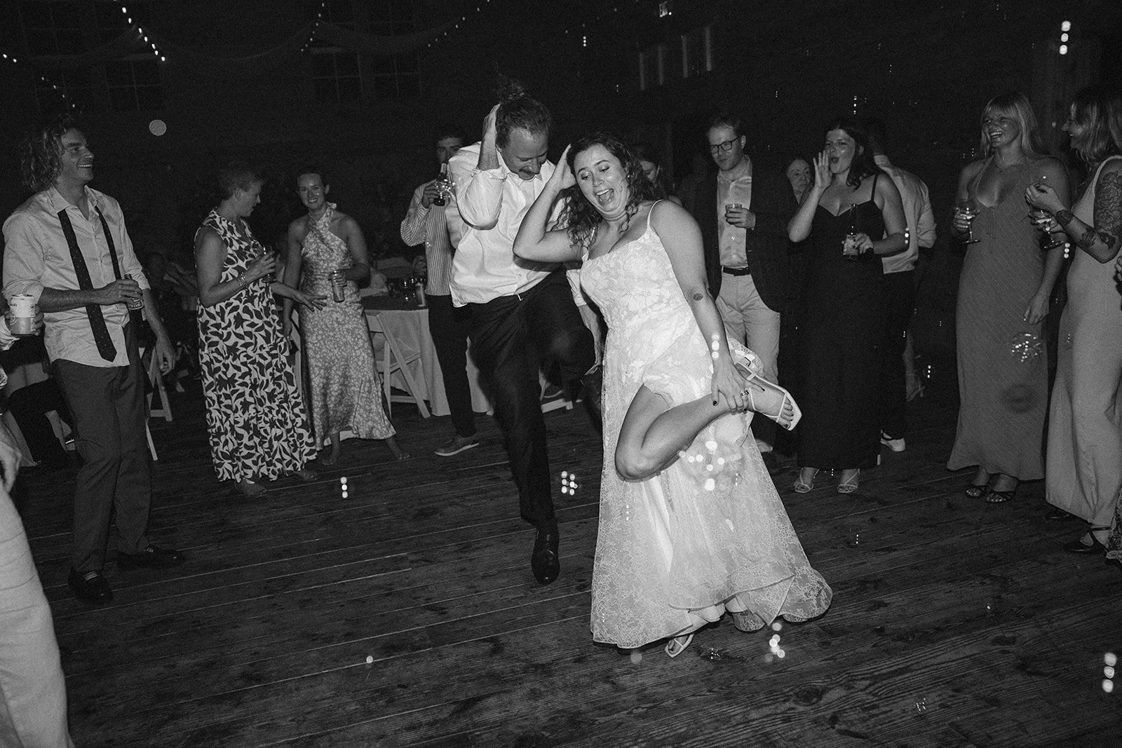 People dancing at a wedding reception, with a bride and groom in the center, surrounded by guests in formal attire, on a wooden dance floor in a dimly lit venue. ocracoke outerbanks wedding film photographer