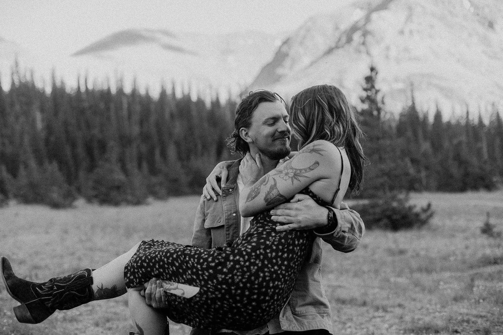 A man is holding a woman in his arms outdoors in a natural setting with trees and mountains in the background. The woman has tattoos on her arm and is wearing a dress, while the man has a beard and is wearing a jacket. They are looking at each other affectionately.