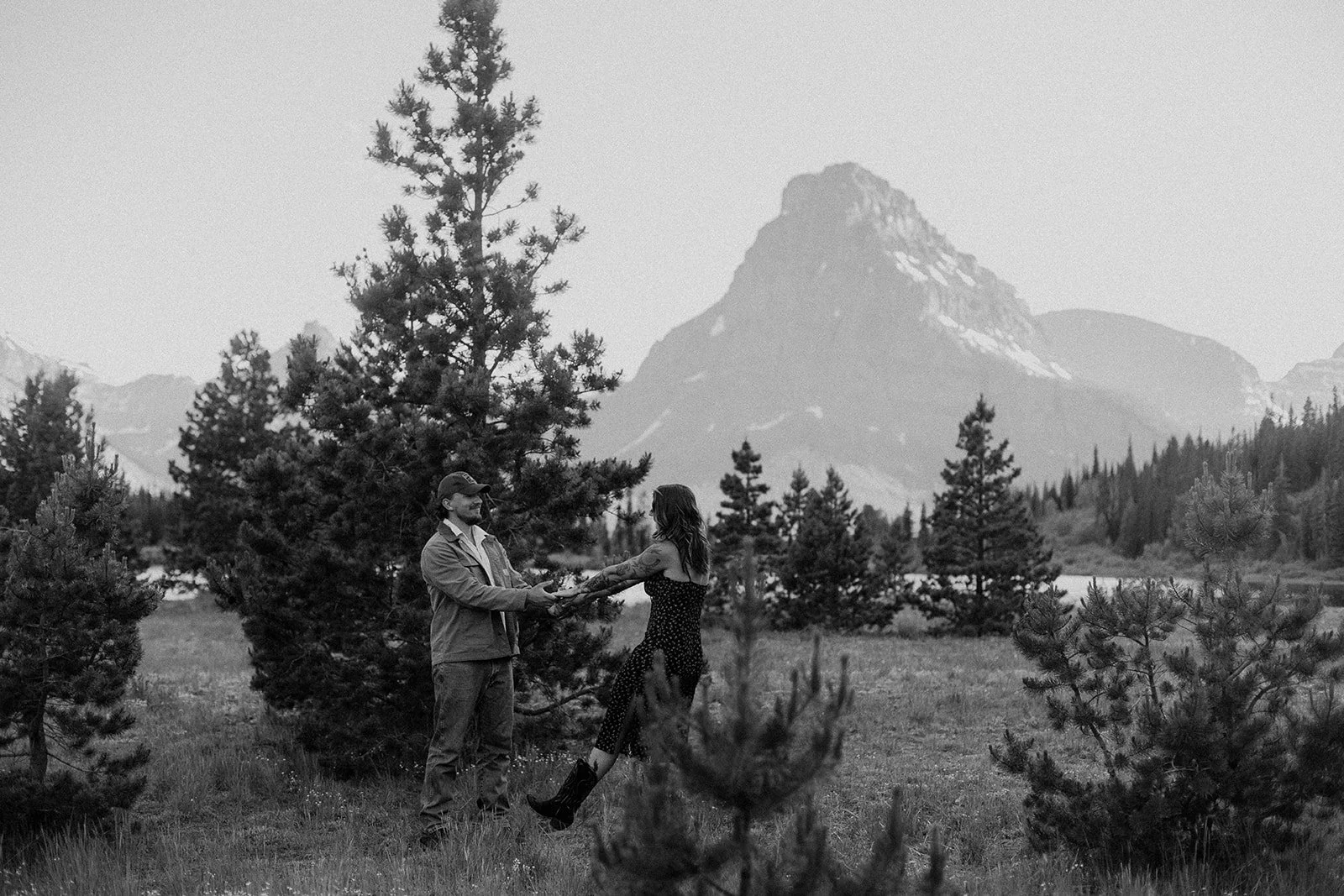 A black and white photo of a couple holding hands in a forested mountain landscape, with a tall mountain in the background, surrounded by trees.