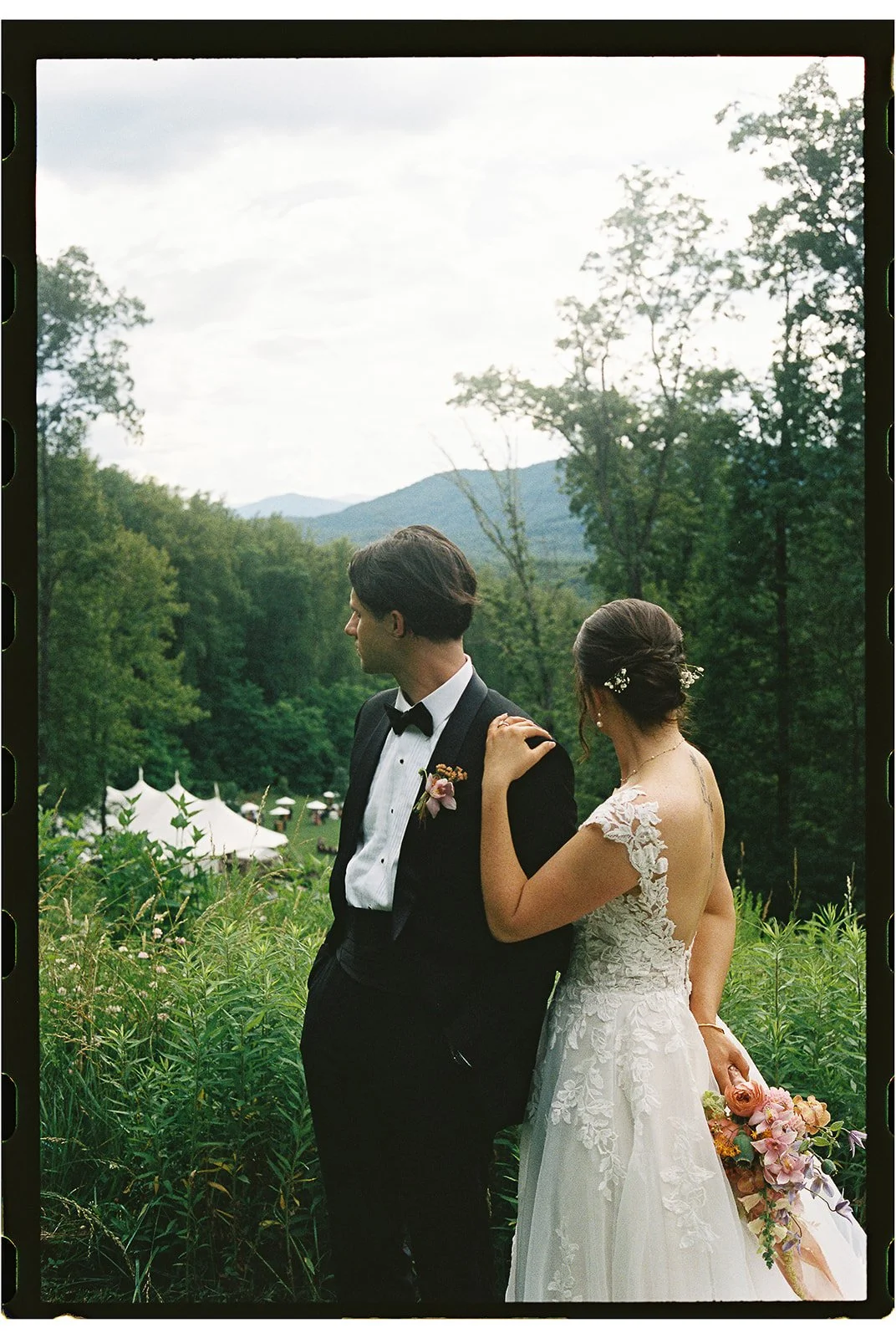A bride and groom standing outdoors among greenery, with a mountain landscape and tents in the background, during a wedding. charlottesville virginia wedding photographer eastwood winery