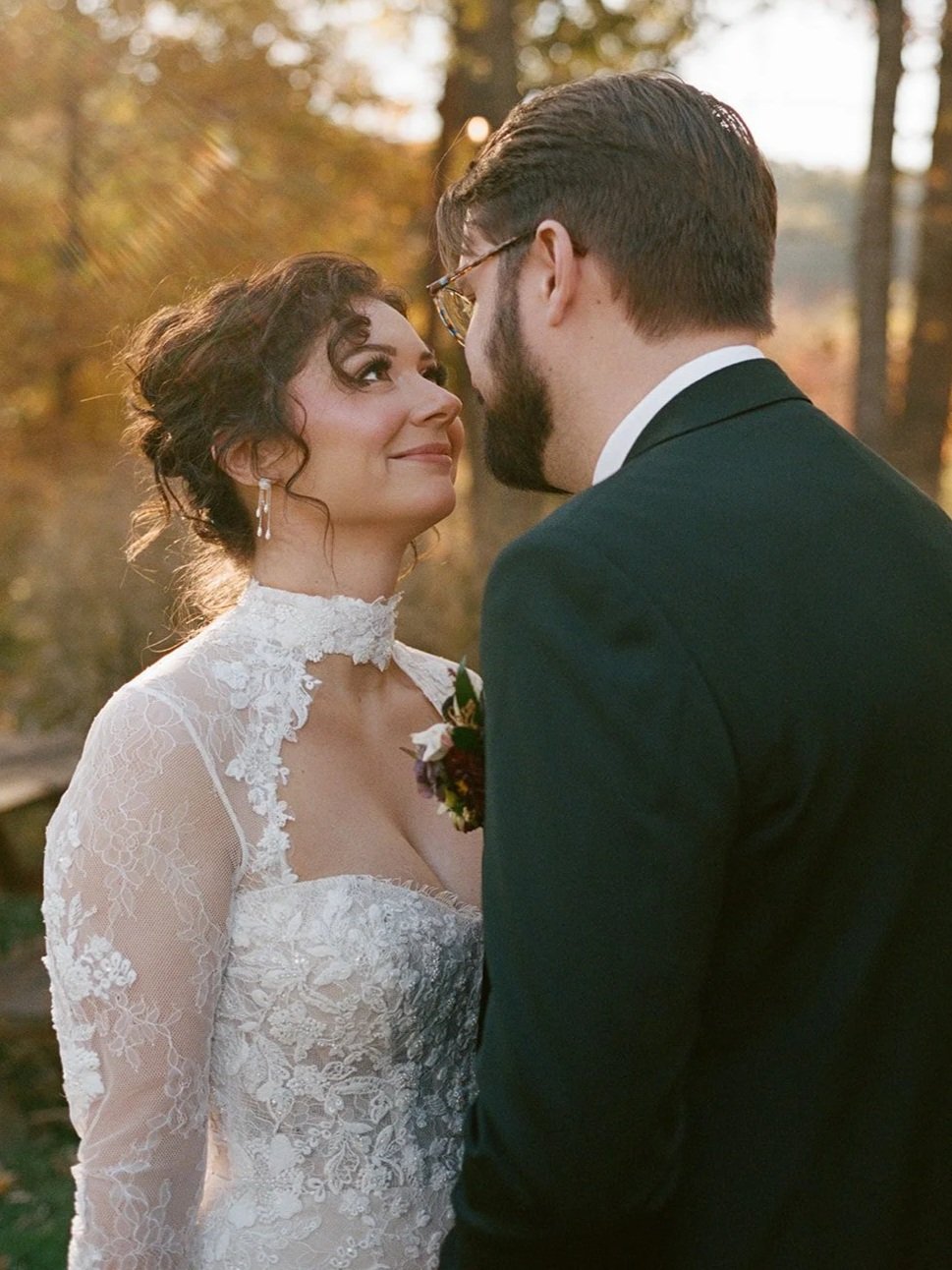 A bride and groom standing close outdoors during sunset, looking into each other's eyes. The bride wears a lace wedding dress with a high neckline and floral details, and the groom wears a black tuxedo with glasses and a beard. potters craft cider