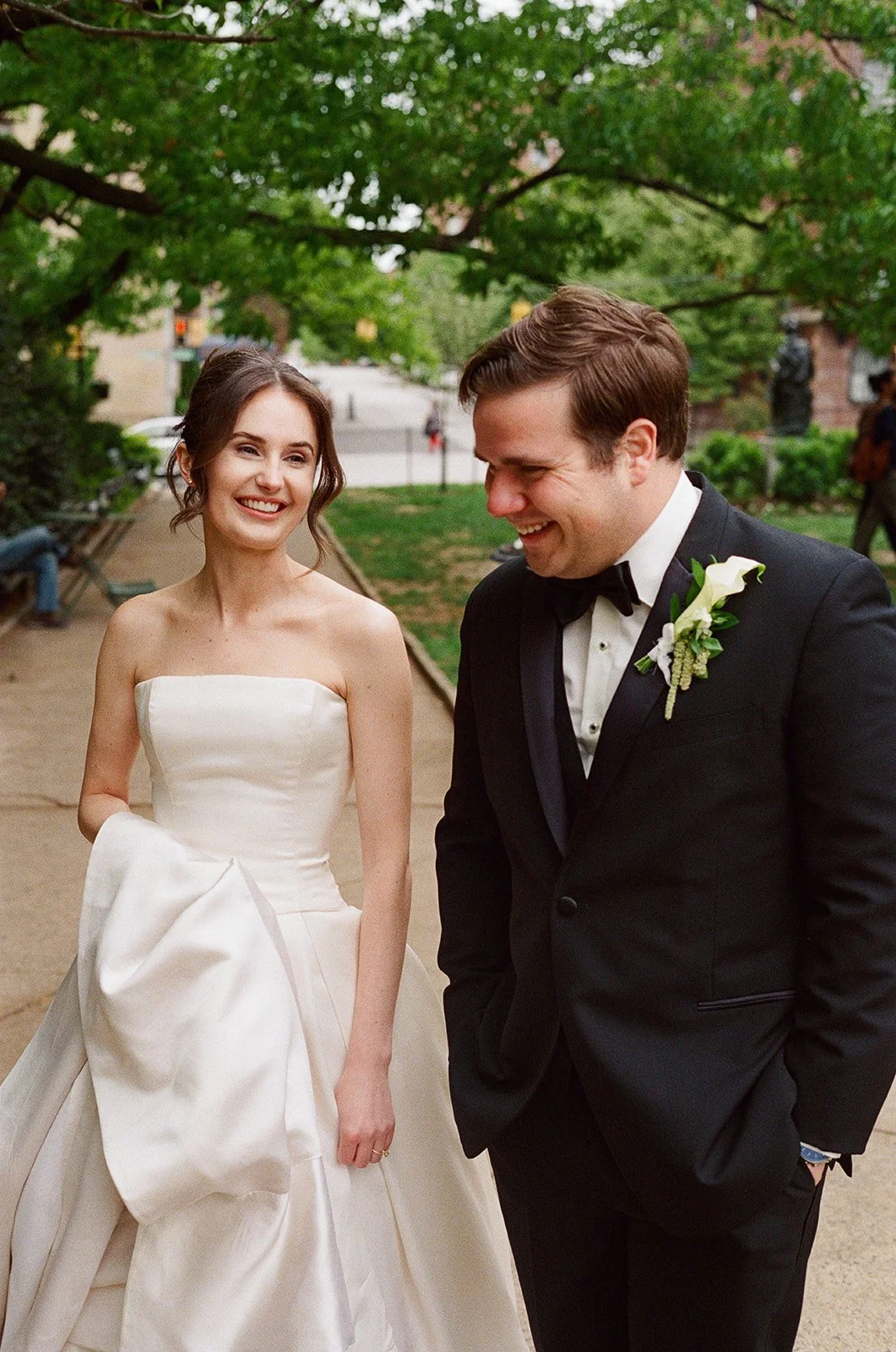 A bride and groom are smiling and laughing outdoors on a wedding day. The bride is wearing a strapless white wedding gown and holding her dress, while the groom is in a black tuxedo with a white boutonniere.