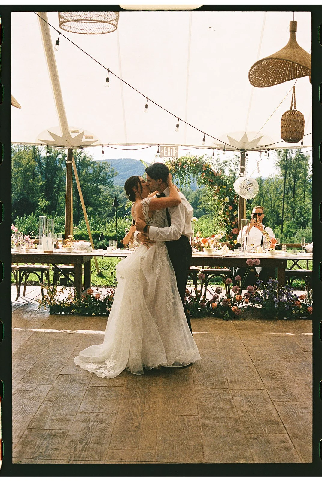A bride and groom share a kiss at their wedding reception under a tent with hanging woven lamps and string lights, surrounded by greenery and flowers, with a man taking a photo in the background. charlottesville virginia wedding photographer eastwood