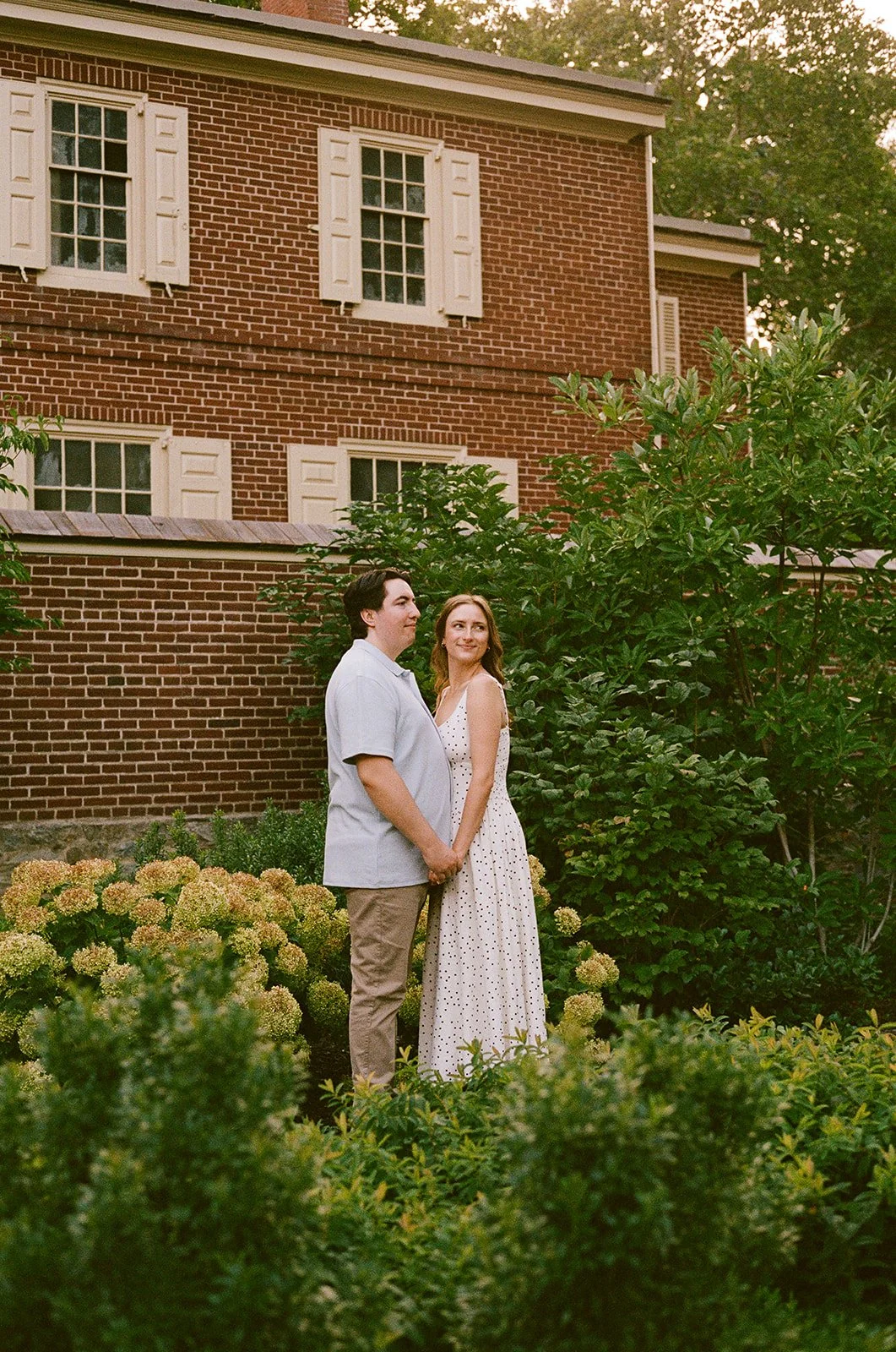 A couple standing hand in hand in a lush garden with yellow flowers, in front of a brick house with white shutters. Old City Philadelphia engagement shoot