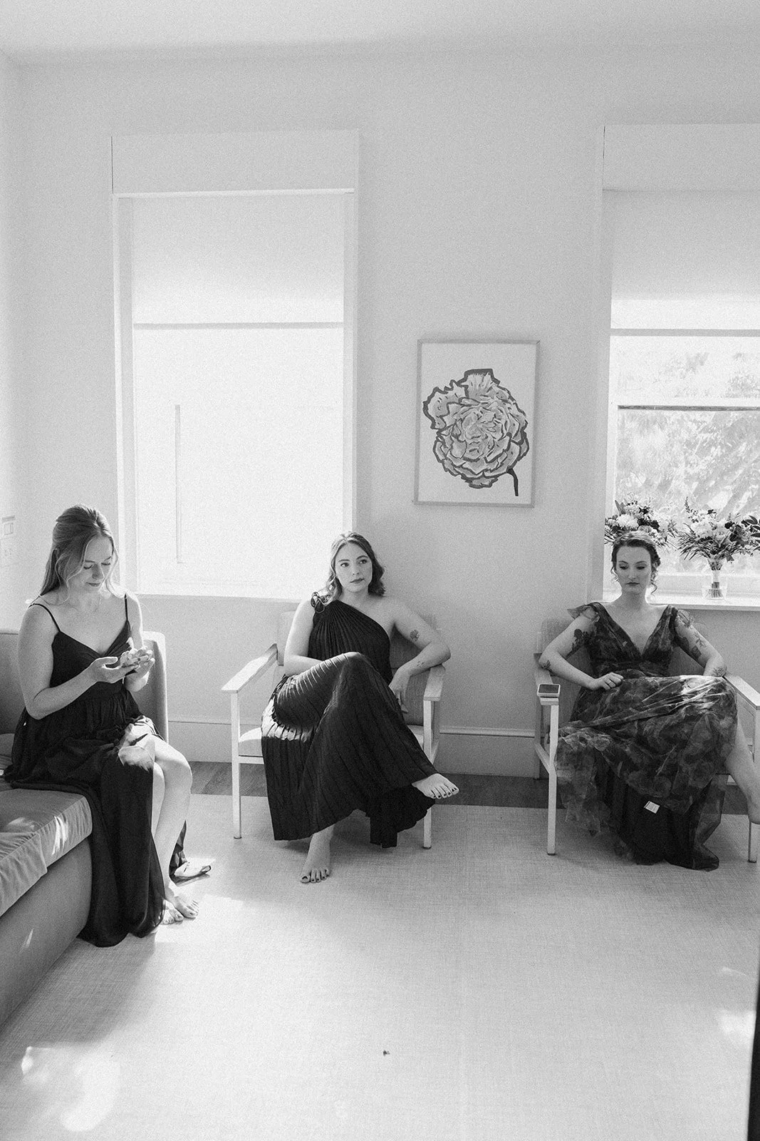 Three women in elegant dresses seated on chairs in a bright room with large windows. One woman is using her phone, and two are sitting with their legs crossed, looking away from each other. doyle hotel charlottesville va