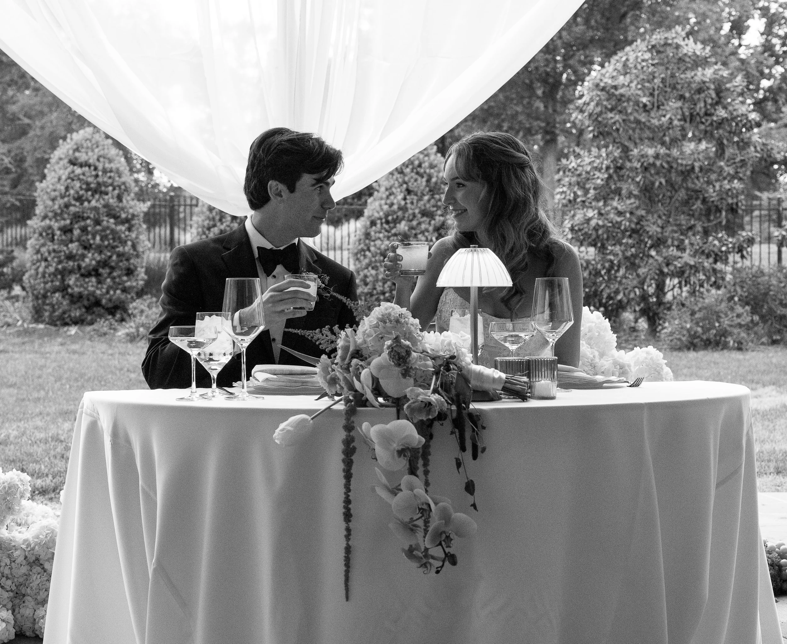 A black and white photo of a bride and groom at a wedding reception, seated at a table with flower arrangements, glasses, and a lamp. They are smiling at each other and raising glasses for a toast outdoors with trees and a fence in the background.