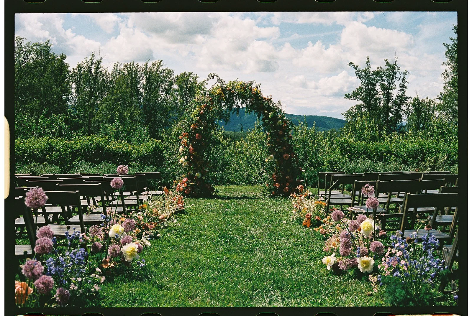 Outdoor wedding setup with a floral arch, chairs arranged on either side, and colorful flower arrangements along the aisle, set in a lush green garden under a partly cloudy sky. charlottesville virginia wedding photographer eastwood winery