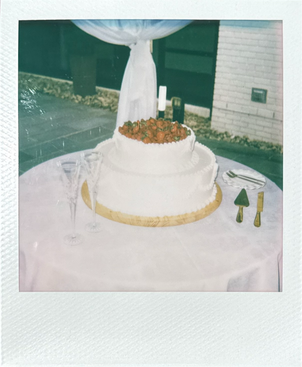 Polaroid photo of a white cake with frosting and strawberries on top, placed on a table with champagne glasses, silverware, and a party hat, outdoors with a brick wall and grass in the background.