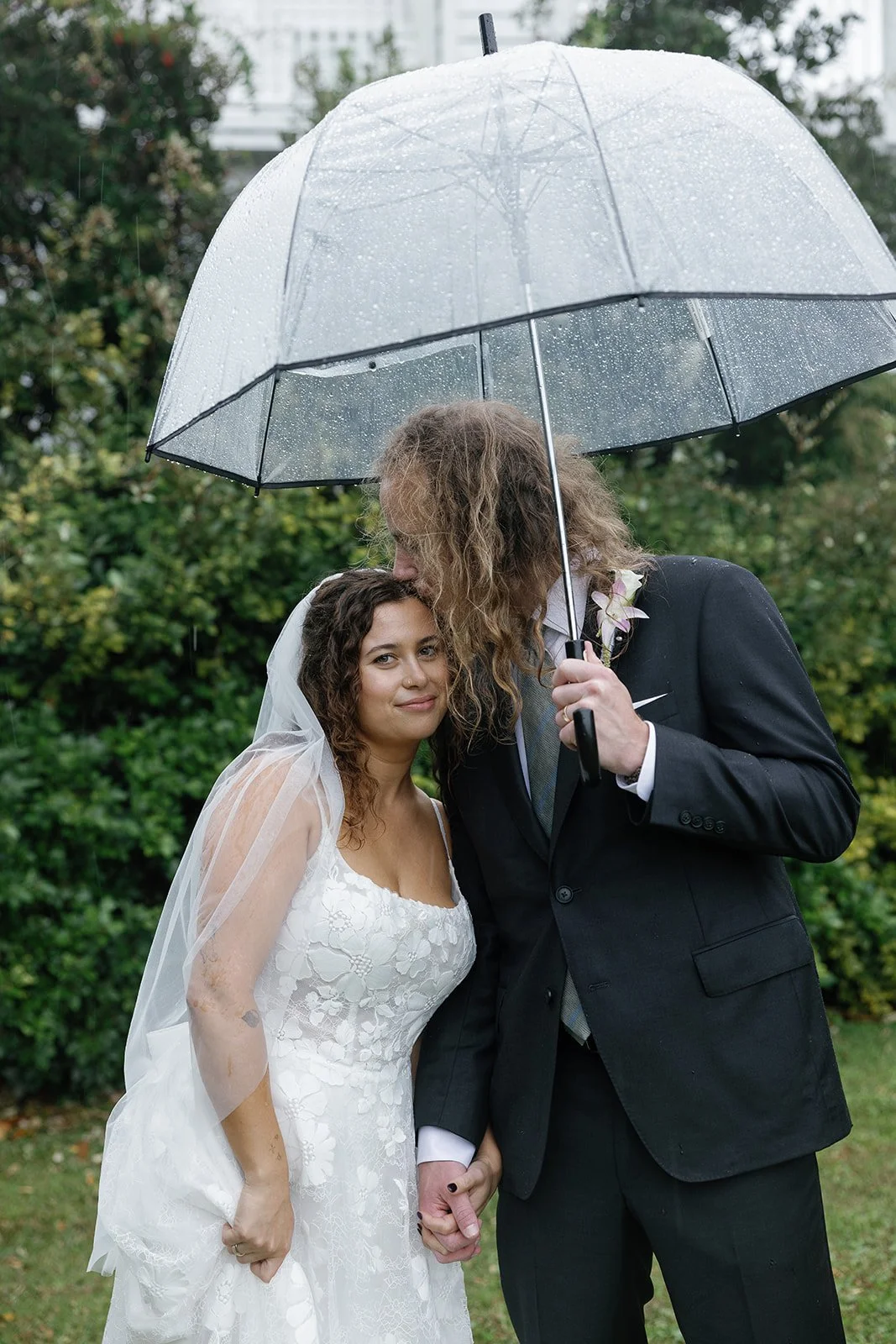 A bride and groom holding hands under a transparent umbrella with raindrops, surrounded by greenery at an outdoor wedding. ocracoke outerbanks wedding film photographer