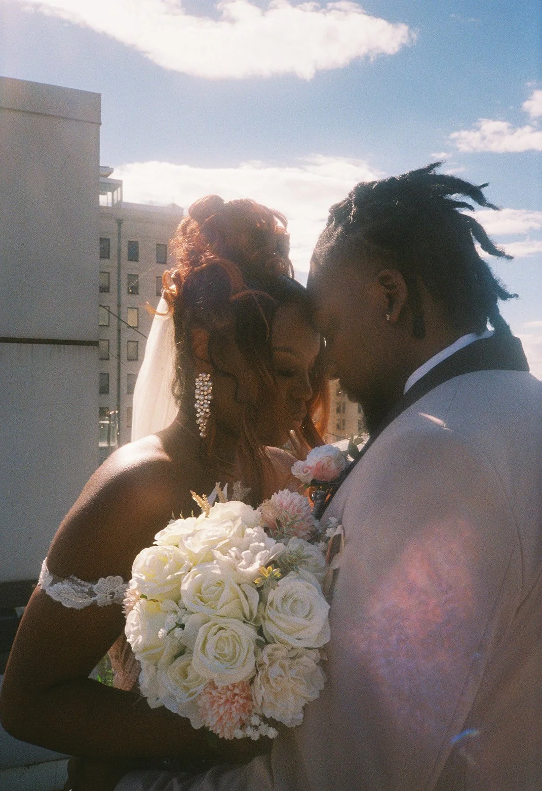 A bride and groom in wedding attire share a gentle, intimate moment outdoors, with the bride holding a bouquet of white and pink roses. roanoke wedding film photographer virginia