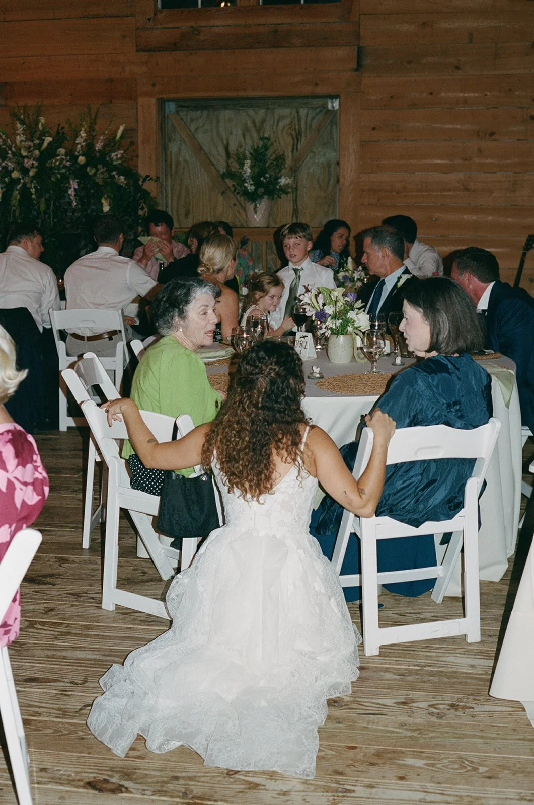 A woman in a white wedding dress kneeling and talking with guests at a wedding reception in a rustic wooden venue. ocracoke outerbanks wedding film photographer