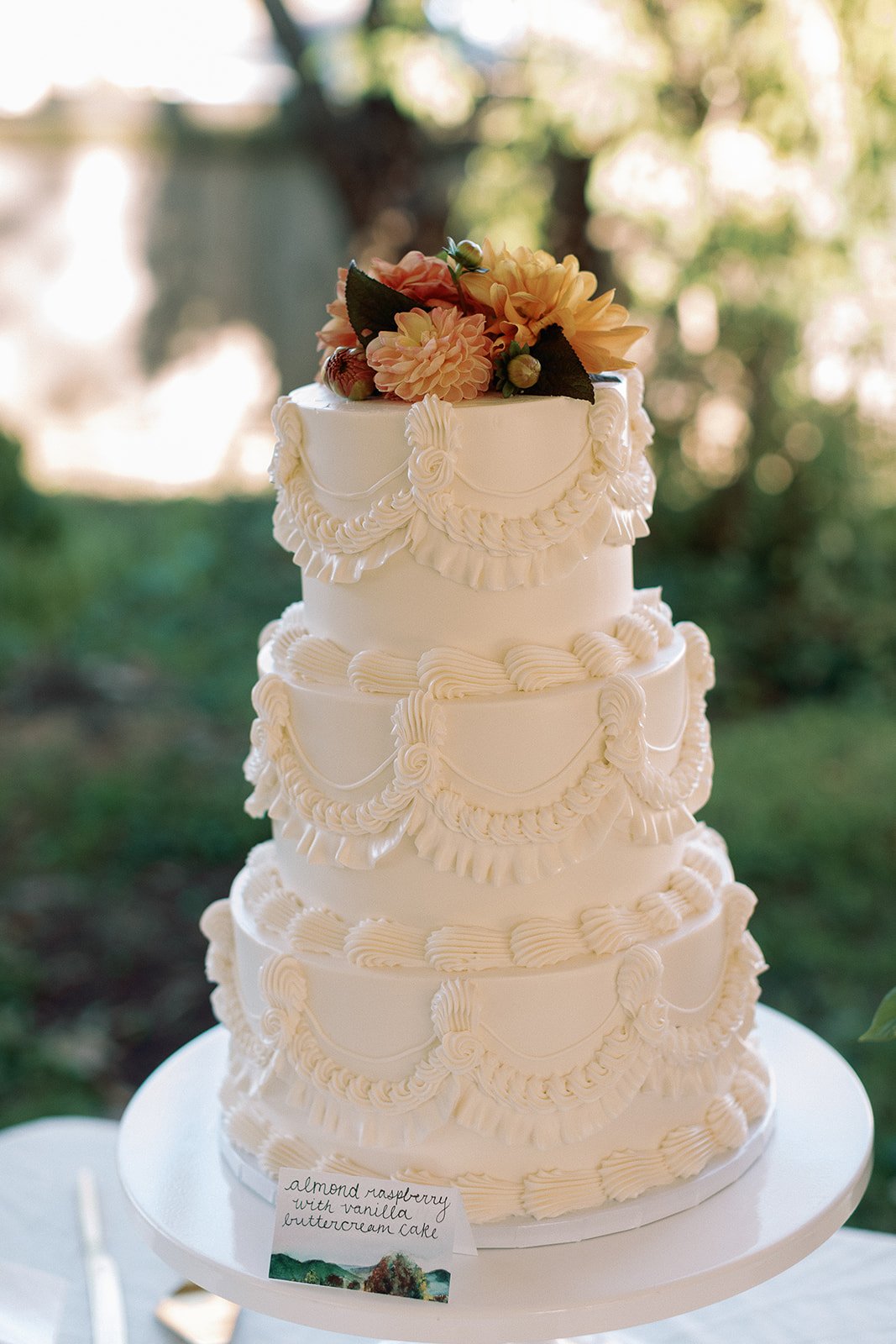 Tall white wedding cake with intricate piping design, topped with a bouquet of peaches, pink roses, berries, and green leaves, on a white table outdoors with a blurred green background. Roanoke Virginia Wedding Film Photographer.