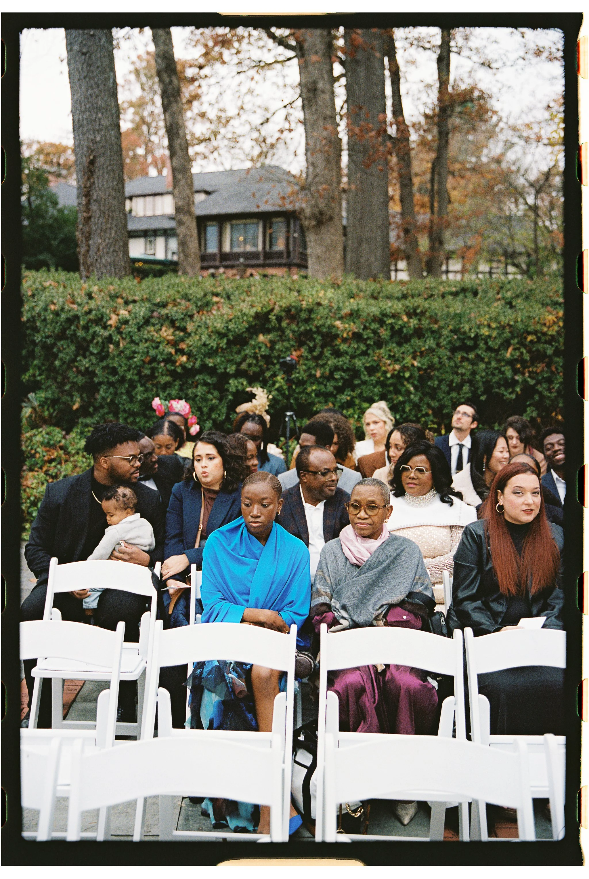 A diverse group of people sitting on white chairs outdoors, dressed in formal attire. The background includes trees and a house, suggesting the setting is a garden or park. Some people are engaged in conversation, while others are looking around. The