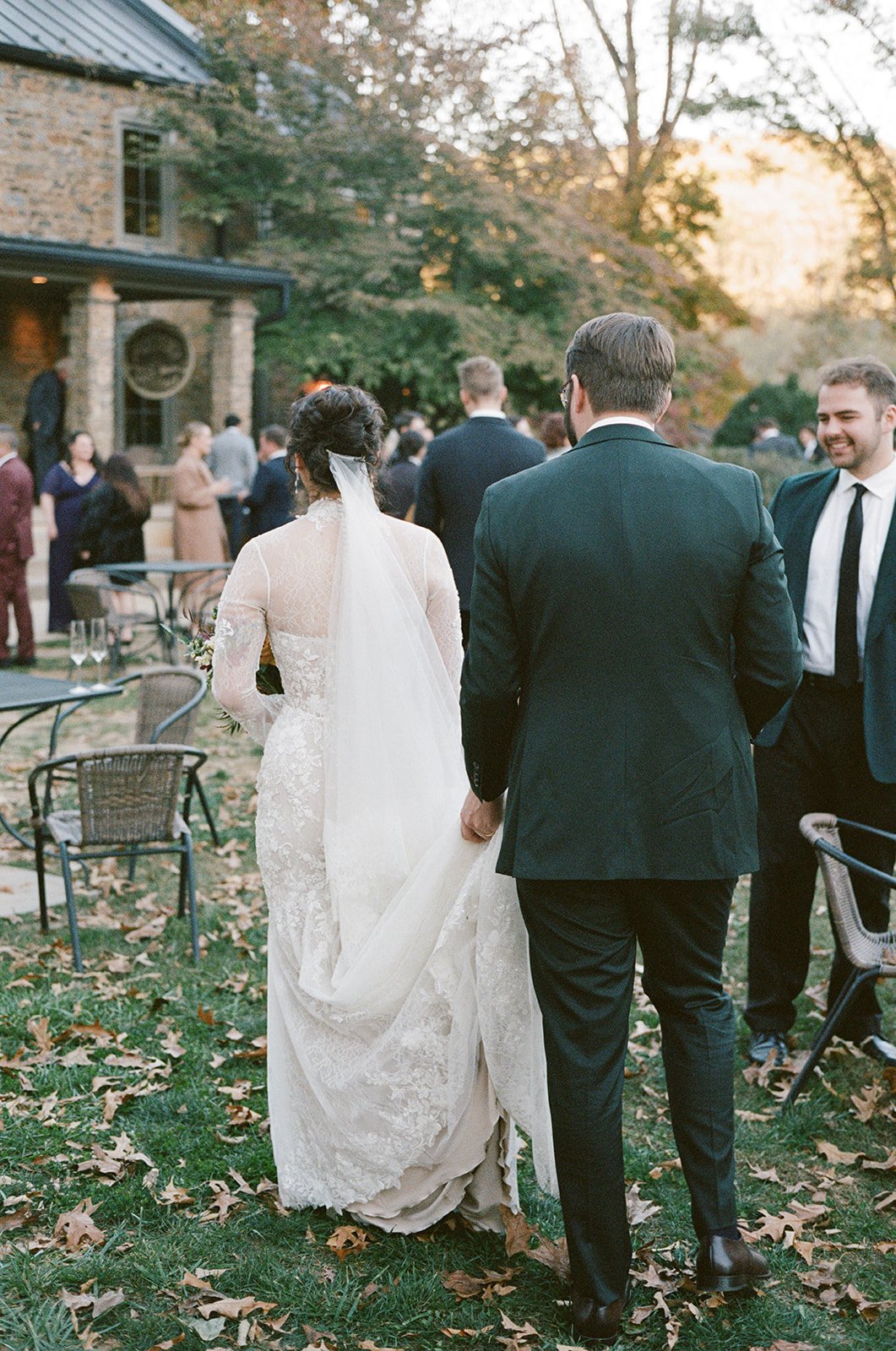 A bride in a long lace wedding dress and veil walking outside with a groom in a dark suit and glasses, while guests gather in the background at an outdoor wedding reception during fall. potters craft cider charlottesville va