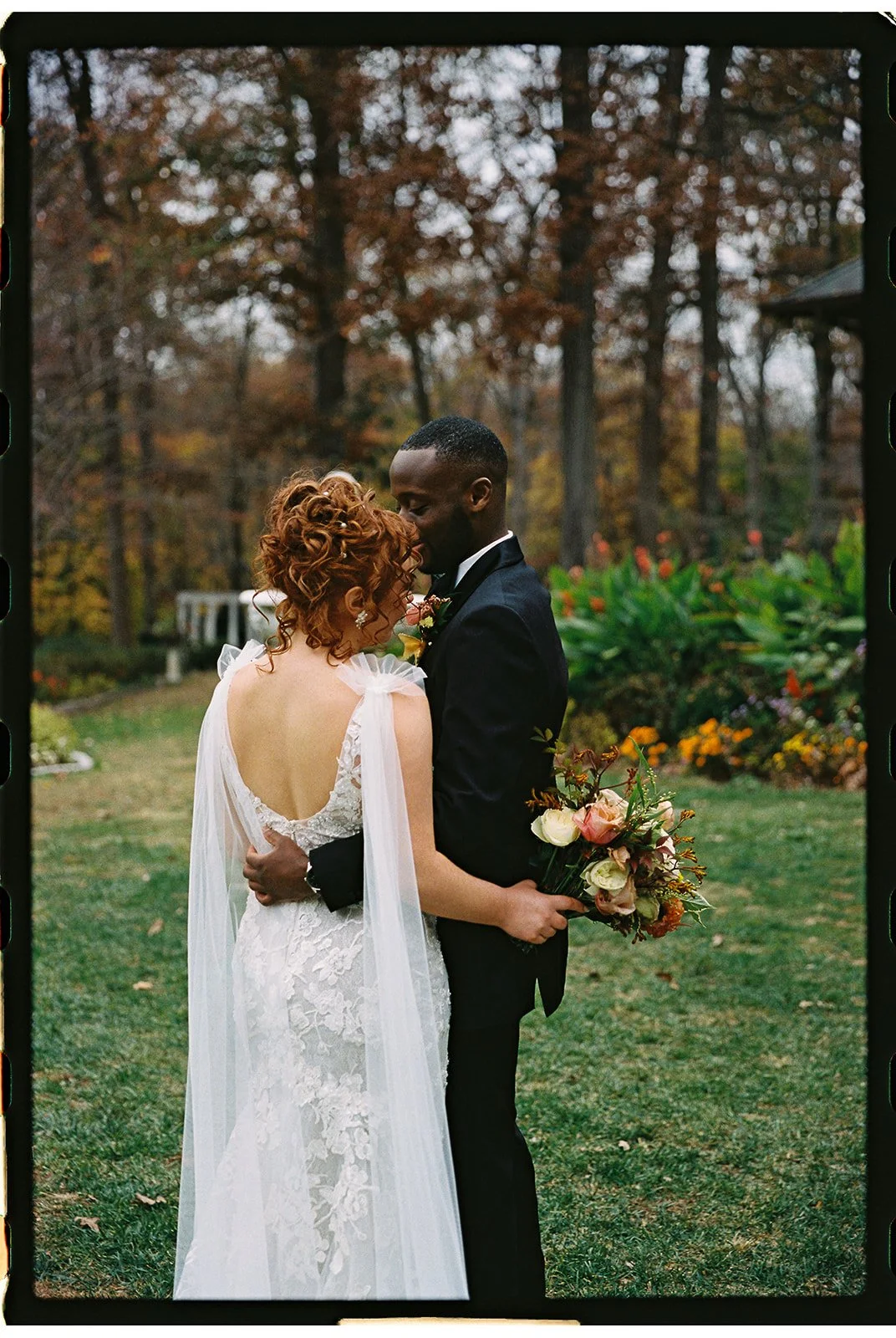 A bride and groom embrace outdoors surrounded by trees and flowers, the bride with curly red hair in a lace wedding dress holding a bouquet, the groom in a black suit. Gramercy Mansion Wedding Film Photographer.