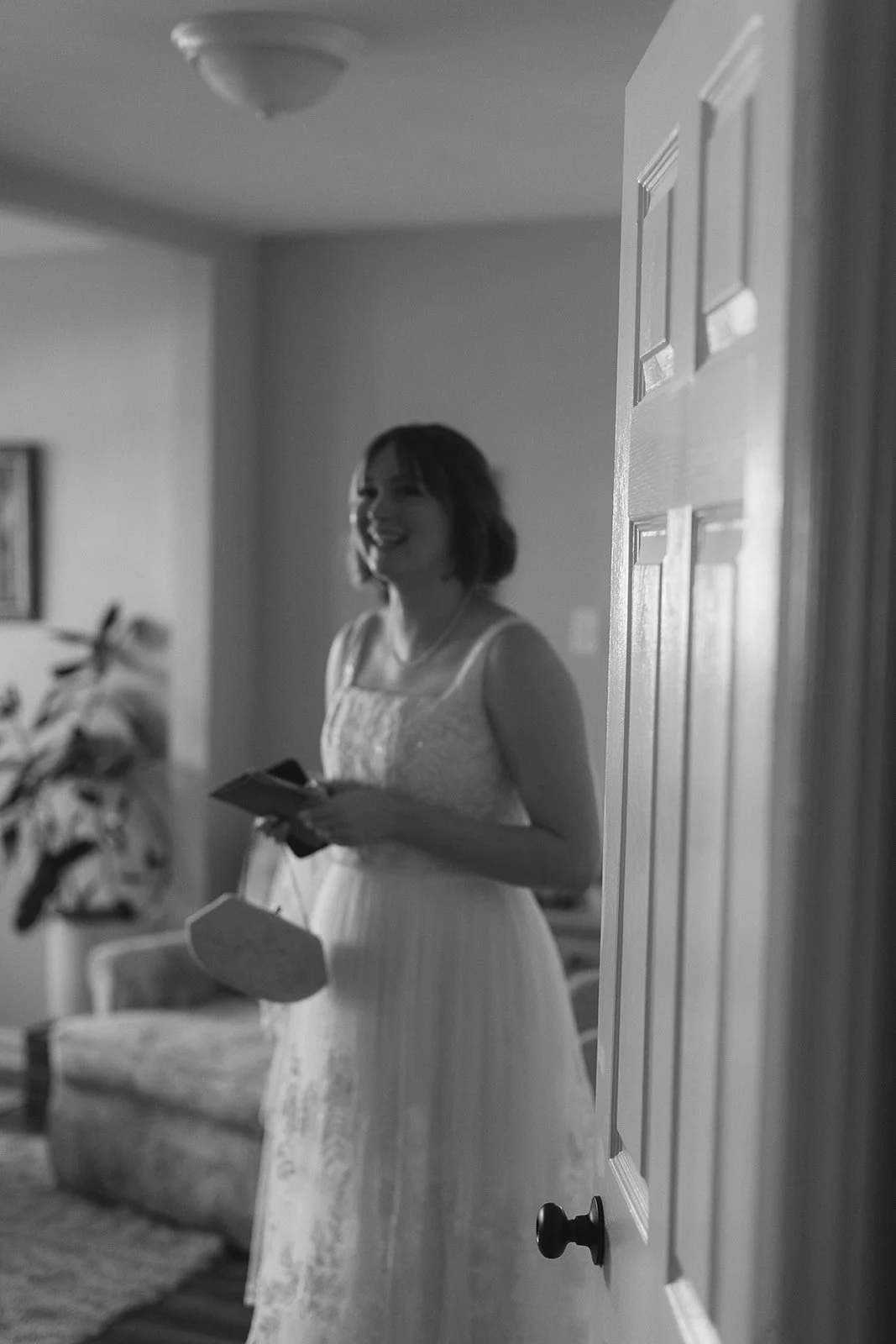 A woman in a white dress smiling and holding a phone, standing in a room with a sofa and a plant in the background. Roanoke Virginia Wedding Photographer.