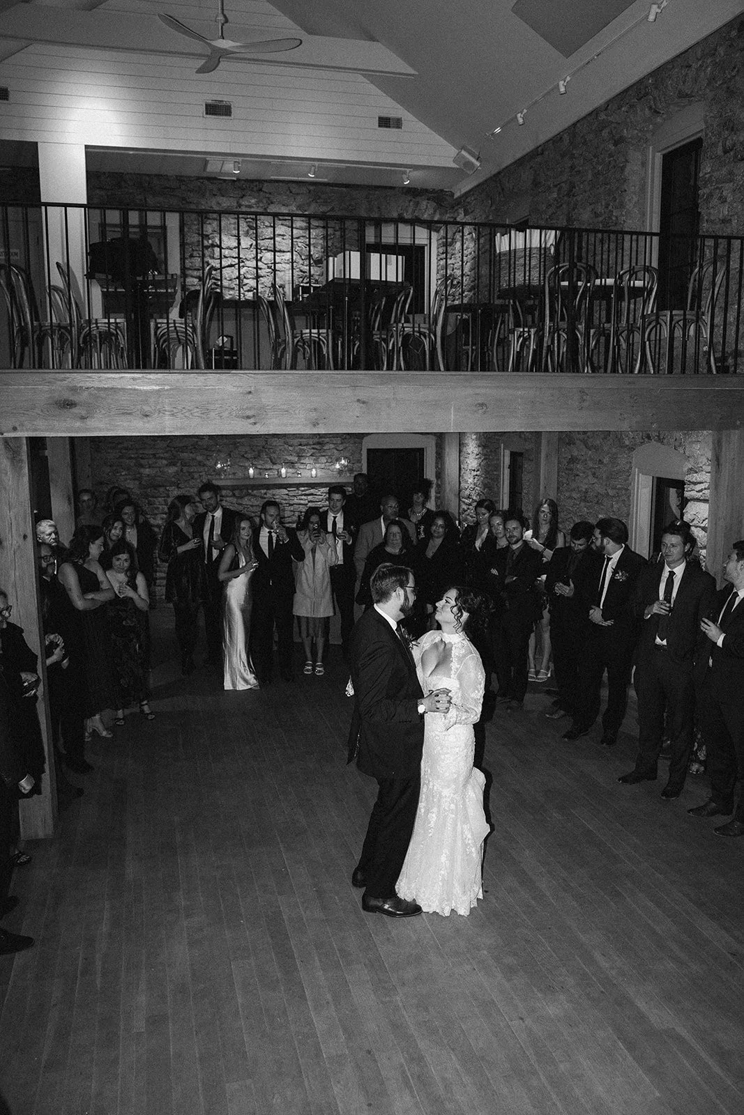 A wedding couple dances in the center of a reception hall with guests watching around them. The hall has brick walls and a wooden floor. potters craft cider charlottesville va