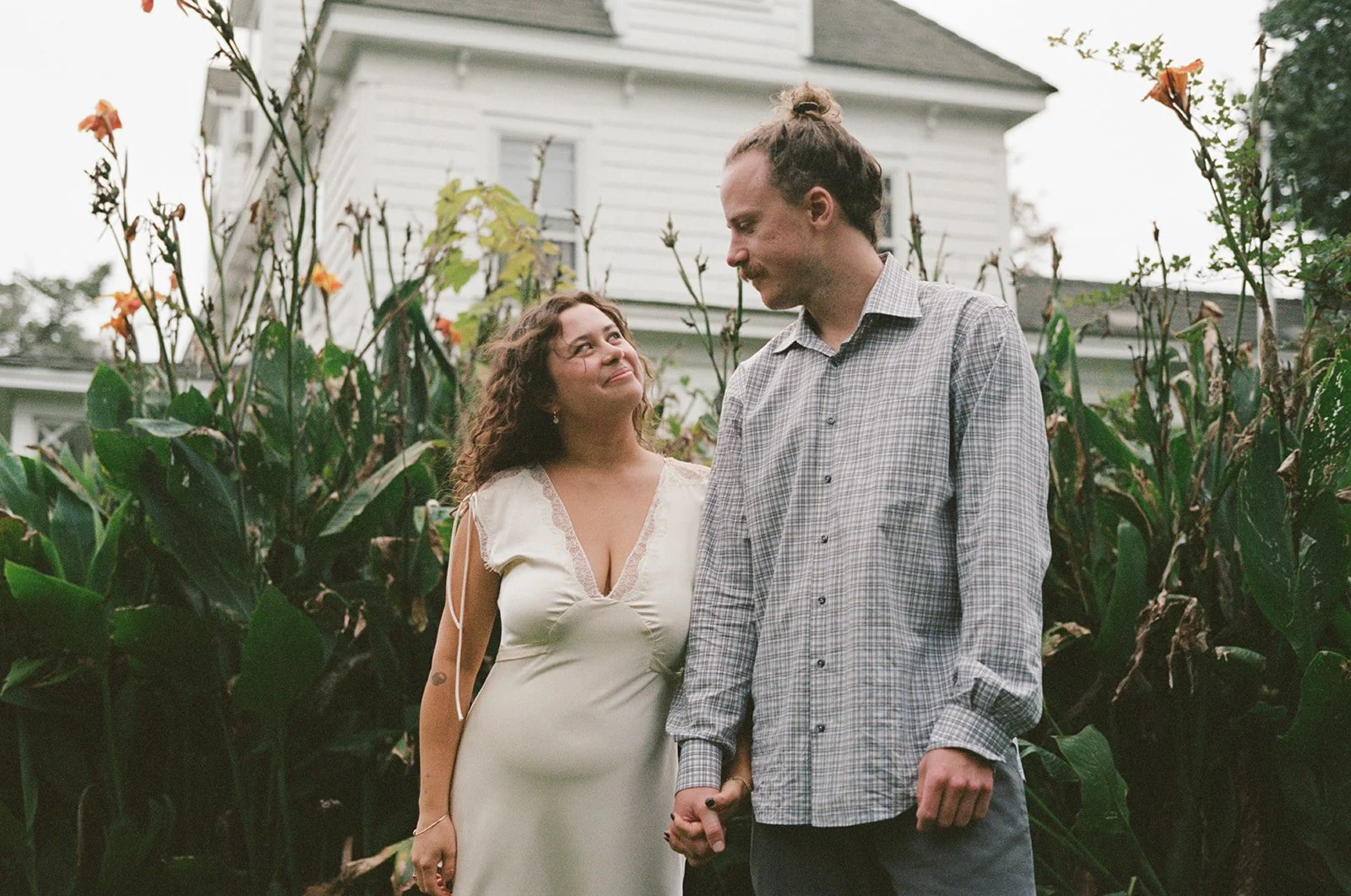 A couple holding hands outdoors in a garden with tall plants, looking at each other. The woman is wearing a cream dress with lace, and the man is wearing a checkered shirt. Behind them is a white house. ocracoke outerbanks wedding film photographer