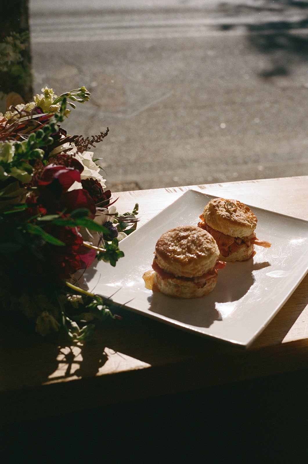 Two sausage biscuits on a white plate beside a flower arrangement in a sunlight-lit setting.