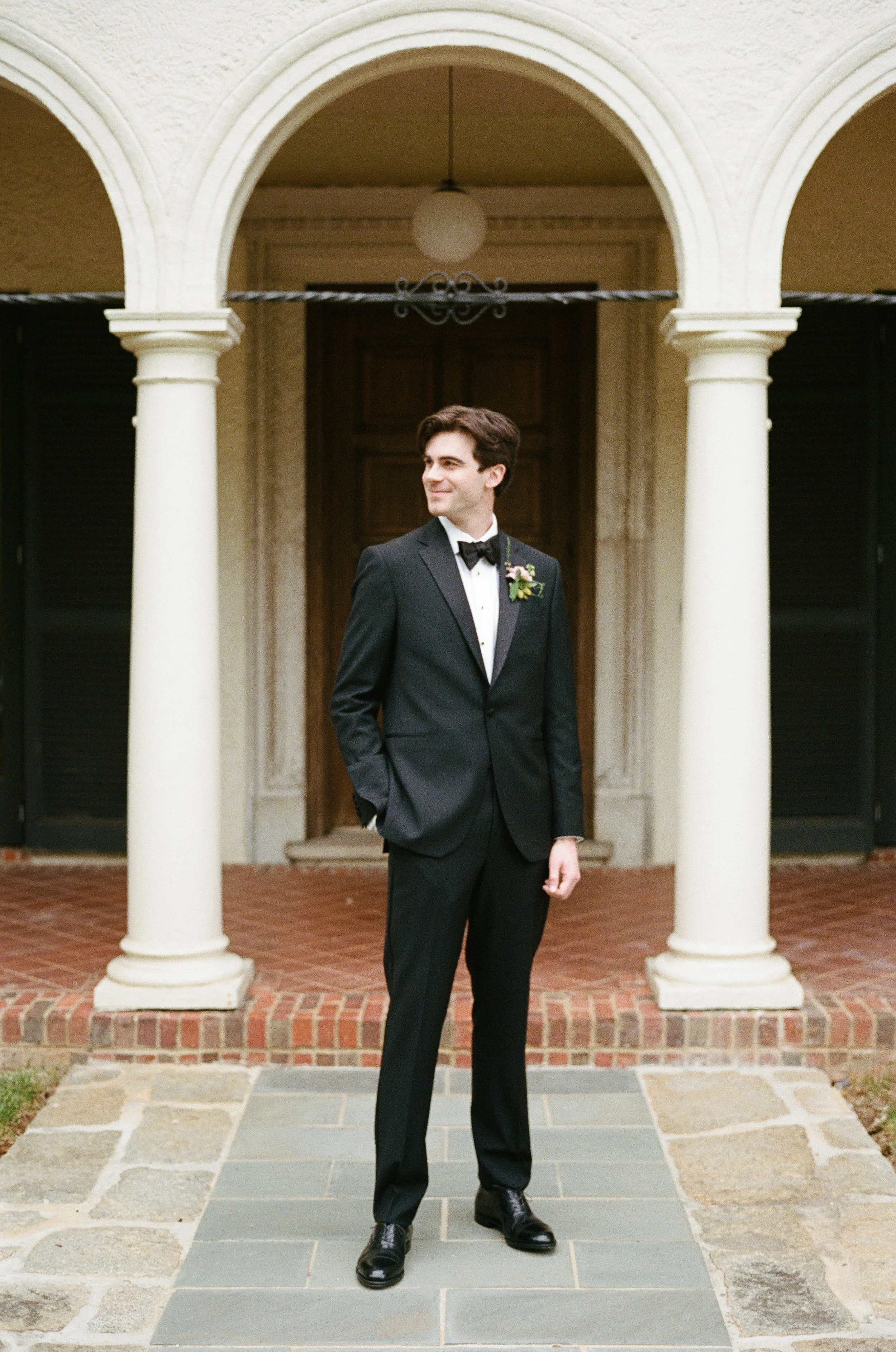 A man in a black tuxedo with a bow tie and boutonniere stands under a white archway on a brick walkway.
