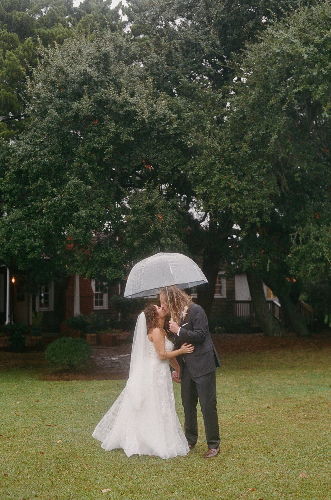 A bride and groom share a kiss under an umbrella on a rainy day, standing on a grassy area with trees and a house in the background. ocracoke outerbanks wedding film photographer