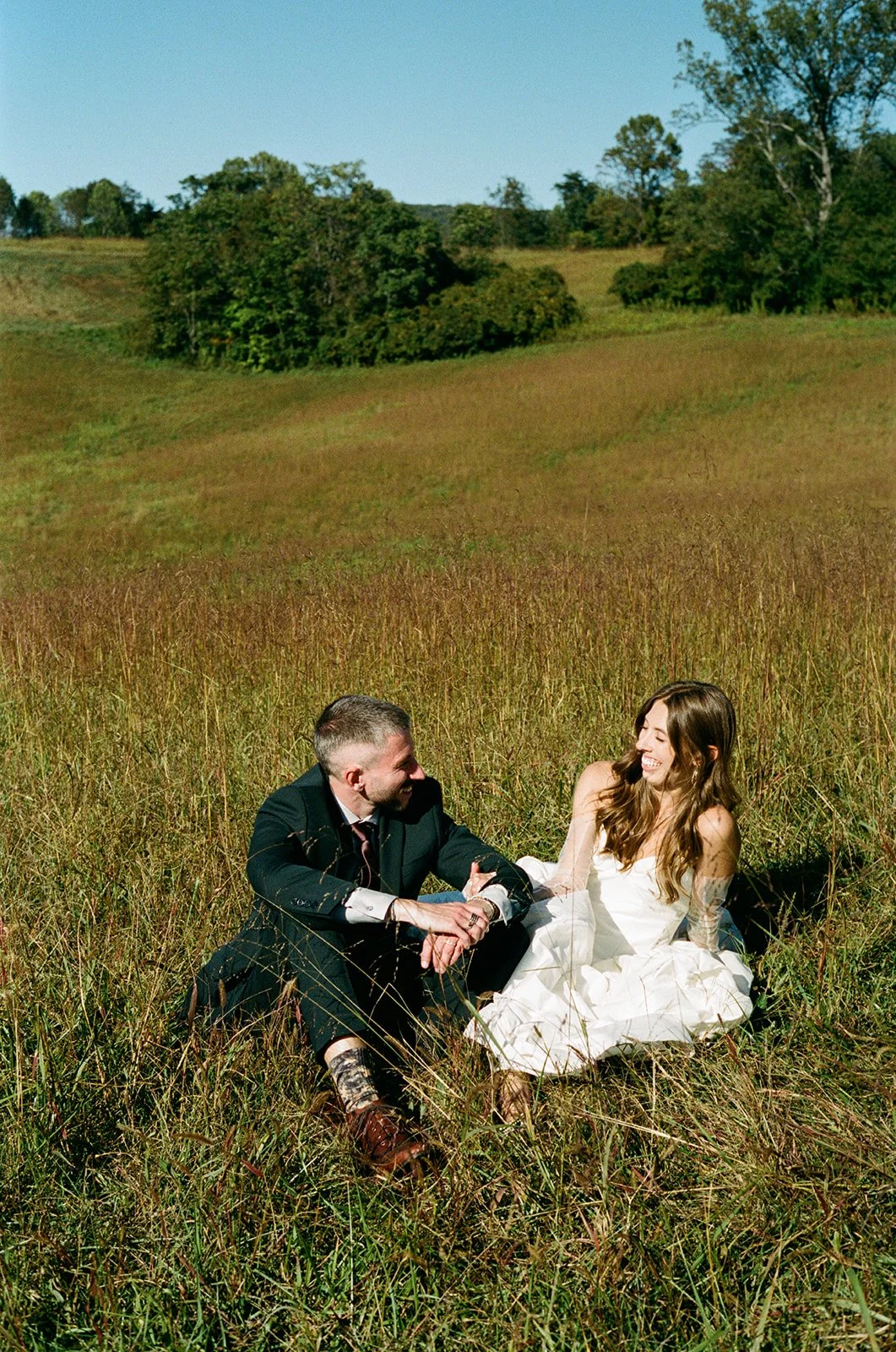 A couple sits in a field of tall grass on a sunny day, smiling and looking at each other. roanoke wedding film photographer virginia