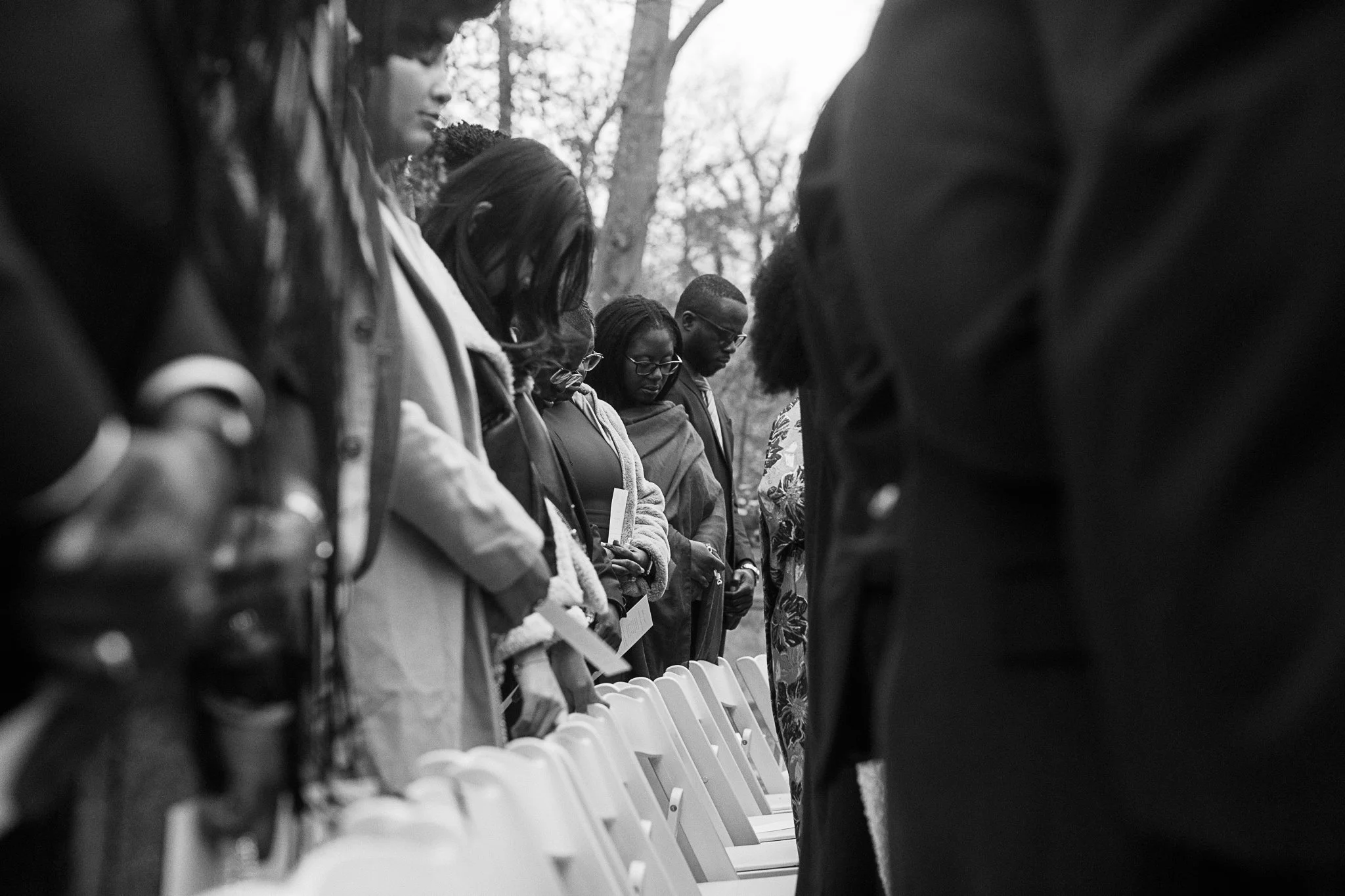 People standing with heads bowed in an outdoor setting, surrounded by empty chairs, wearing formal attire. gramercy mansion maryland wedding