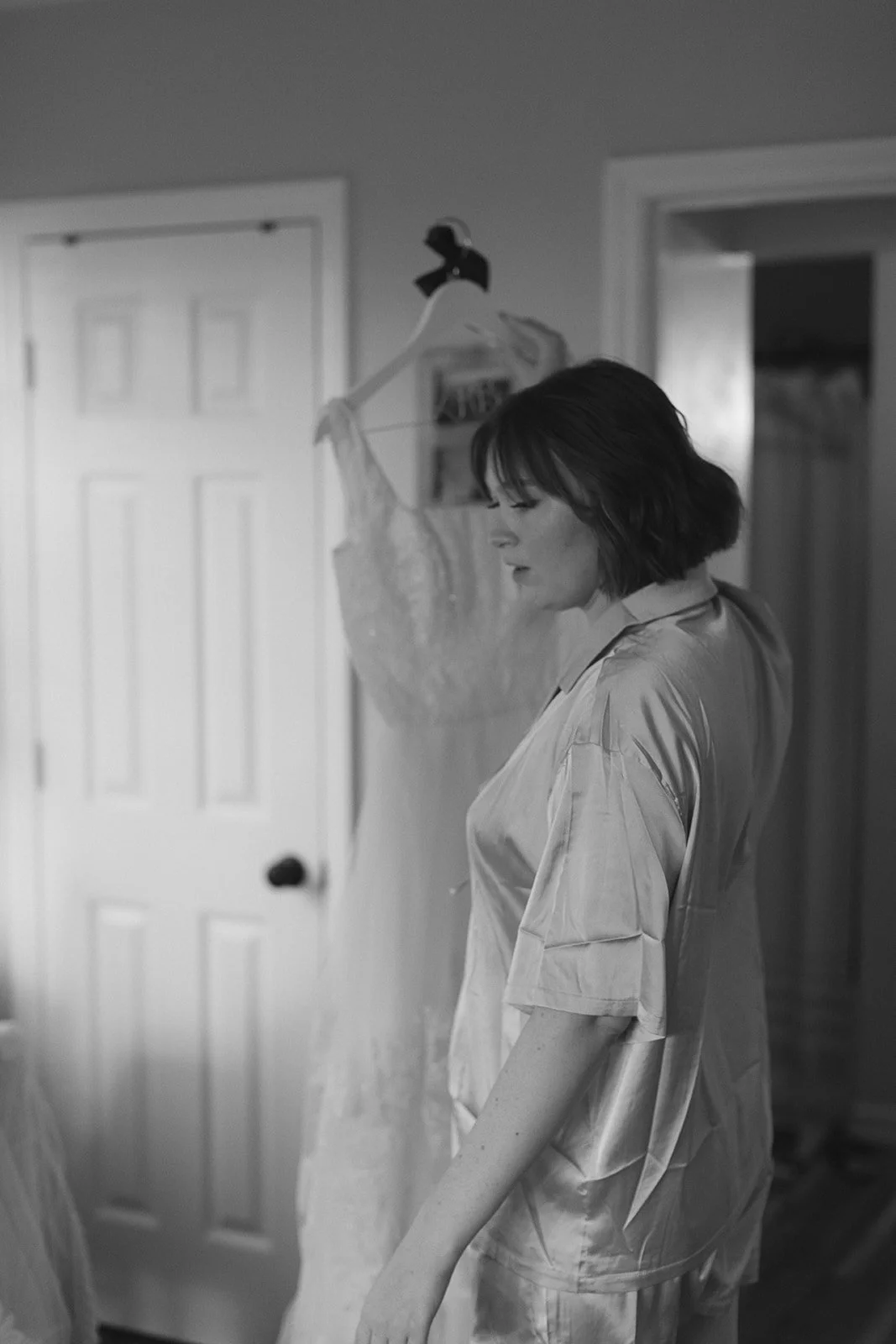 A woman in satin pajamas standing in a bedroom, looking down with a contemplative expression, with a lace dress hanging on a clothes hanger in the background. Roanoke Virginia Wedding Photographer.
