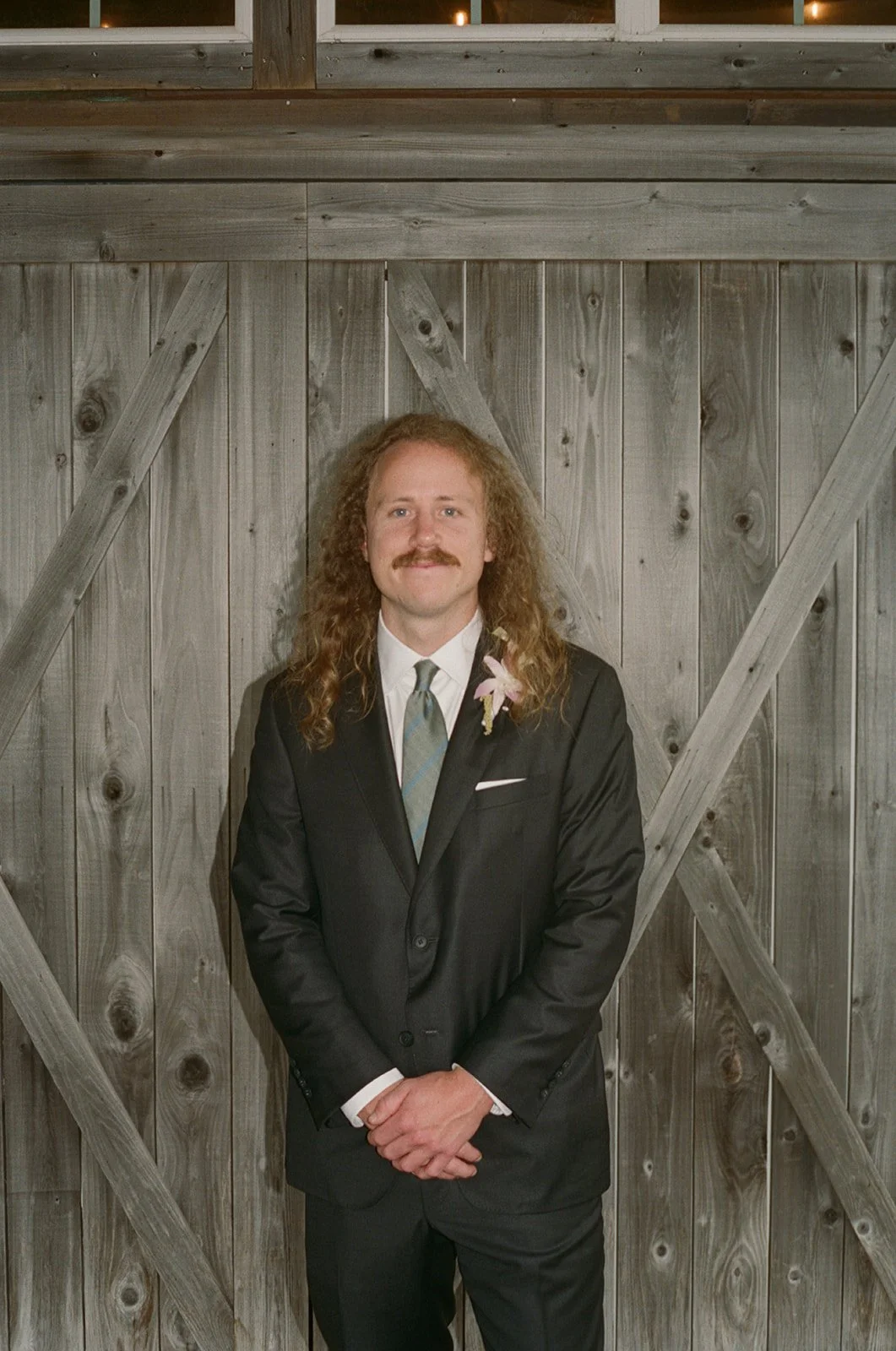 A man with long curly hair and a mustache wearing a black suit, white shirt, and tie standing in front of a wooden rustic background, with a boutonniere on his left lapel. ocracoke outerbanks wedding film photographer