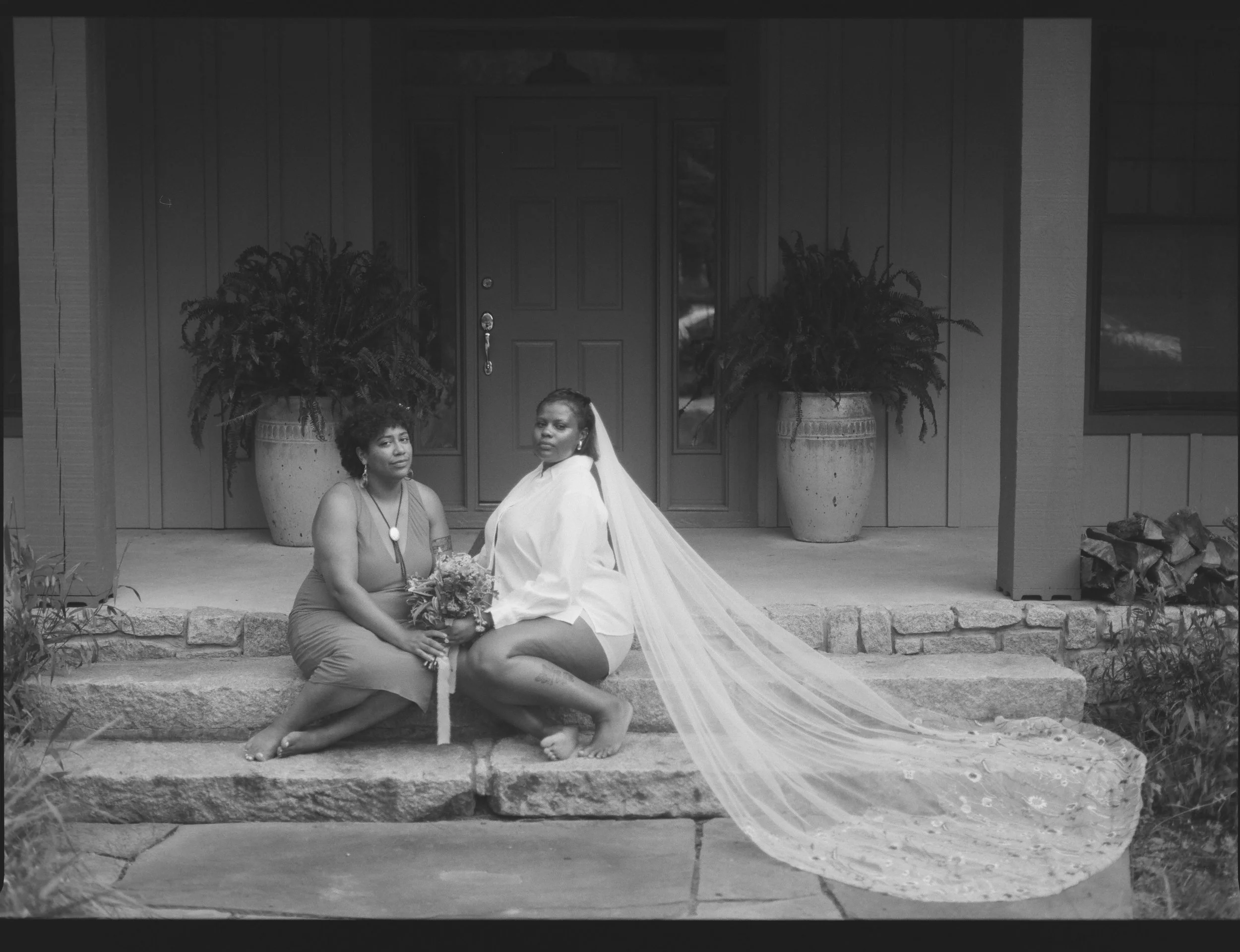 A queer couple sitting on stone steps in front of a house. One is wearing a bridal veil and holding a bouquet, while the other is sitting beside her. Two large potted plants are visible behind them.