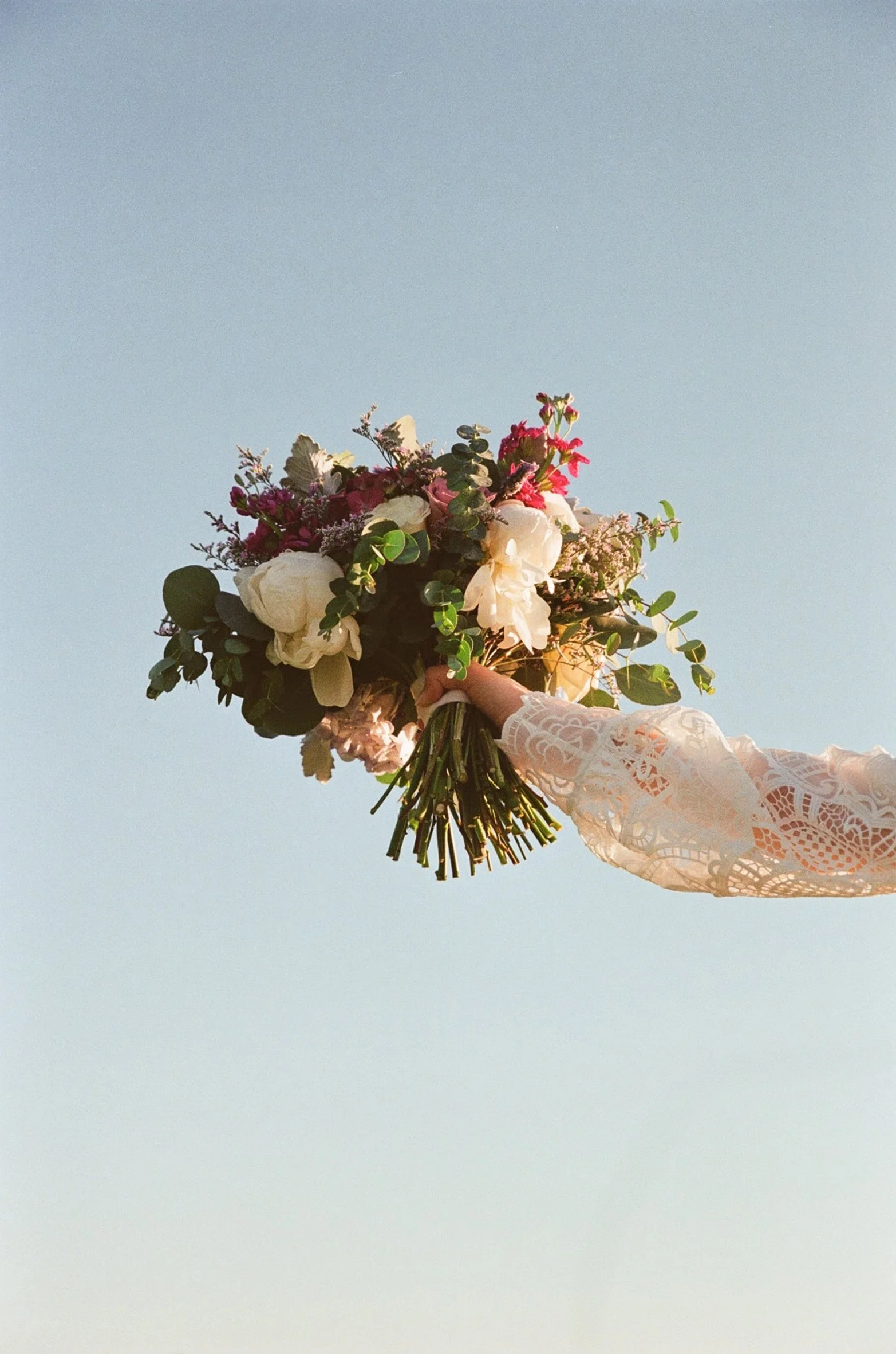 A hand holding a bouquet of mixed flowers, including white roses and greenery, against a clear blue sky.