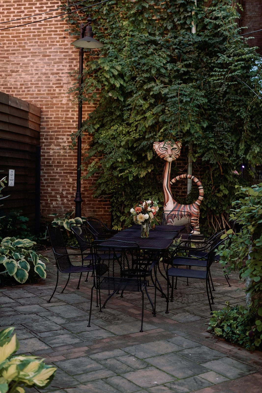 An outdoor patio with a black metal table and six matching chairs, decorated with a vase of pink and white flowers. In the background, a large striped tiger sculpture with a circular tail and a face with cat-like features stands near green ivy-covere