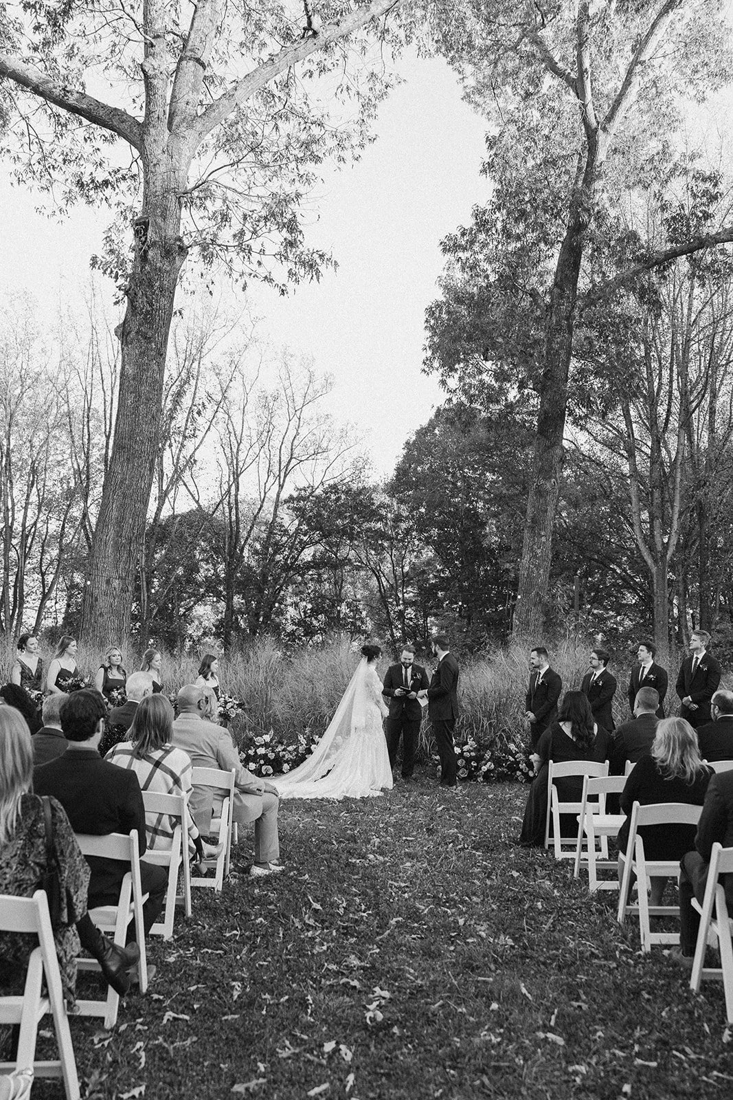 A black-and-white outdoor wedding ceremony with a bride and groom under a tree, surrounded by their wedding party and guests seated on chairs. potters craft cider charlottesville va