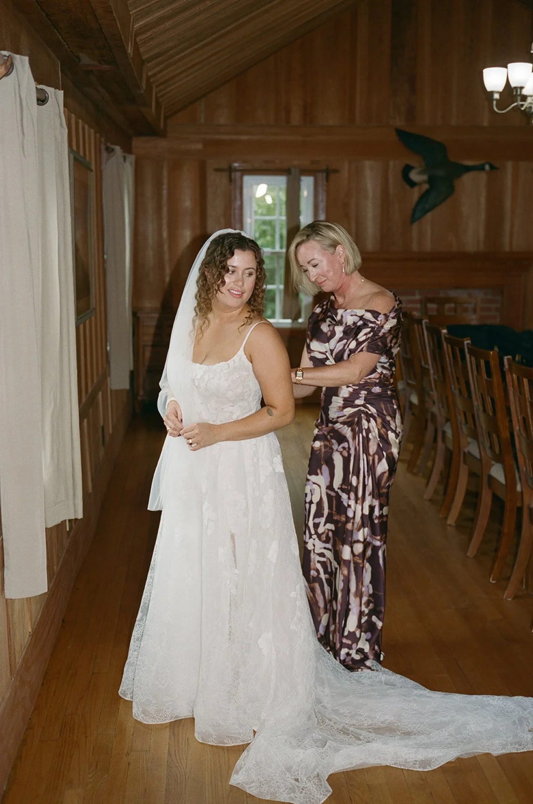 A bride in a white wedding dress and veil getting help with her dress from an older woman in a floral dress, inside a wooden-paneled room with chairs and a bird decoration on the wall. ocracoke wedding north carolina Wedding Film Photographer